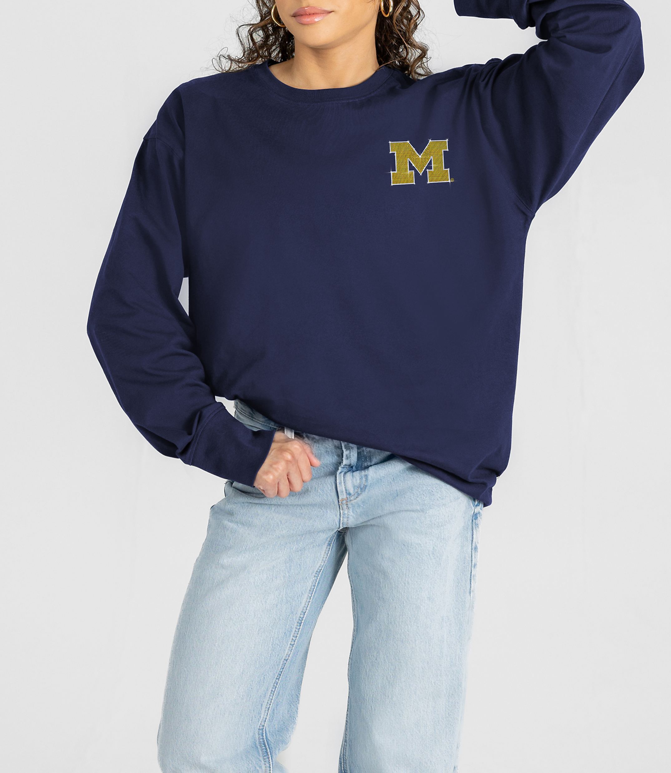 A woman in a navy sweatshirt with a gold "M" logo and light-wash jeans stands against a plain white background.