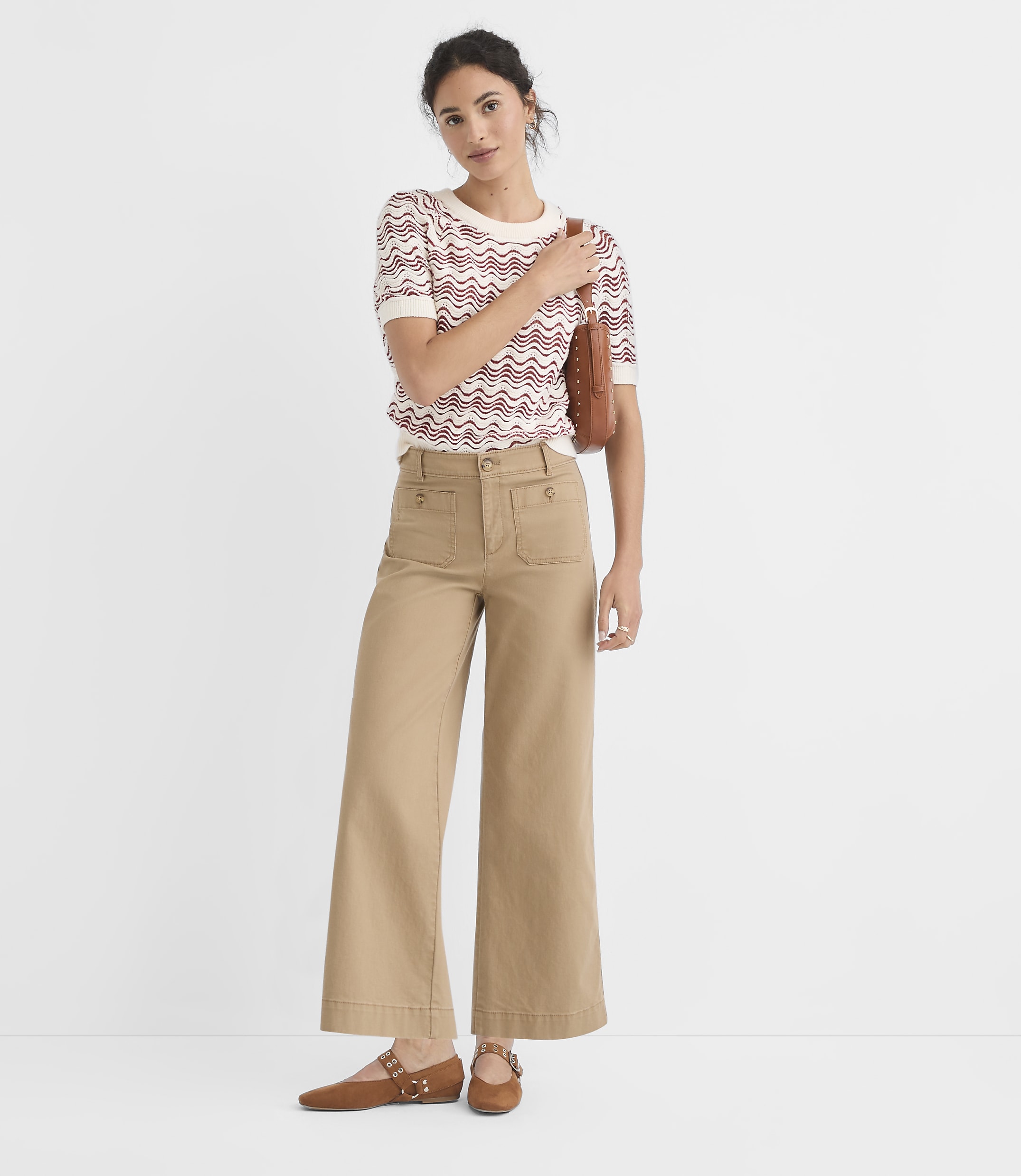 A woman in a patterned sweater and tan pants stands against a plain white background.