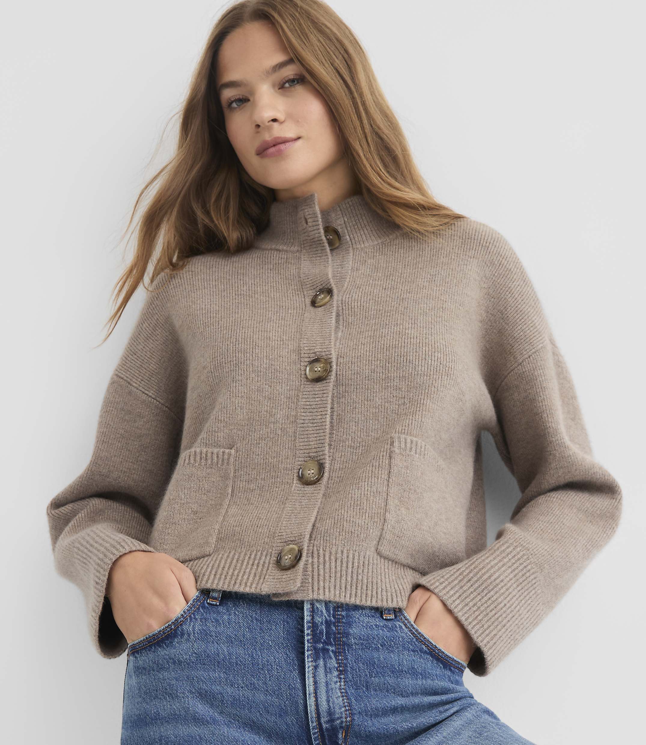 A woman in a beige cardigan and blue jeans stands against a plain white background.