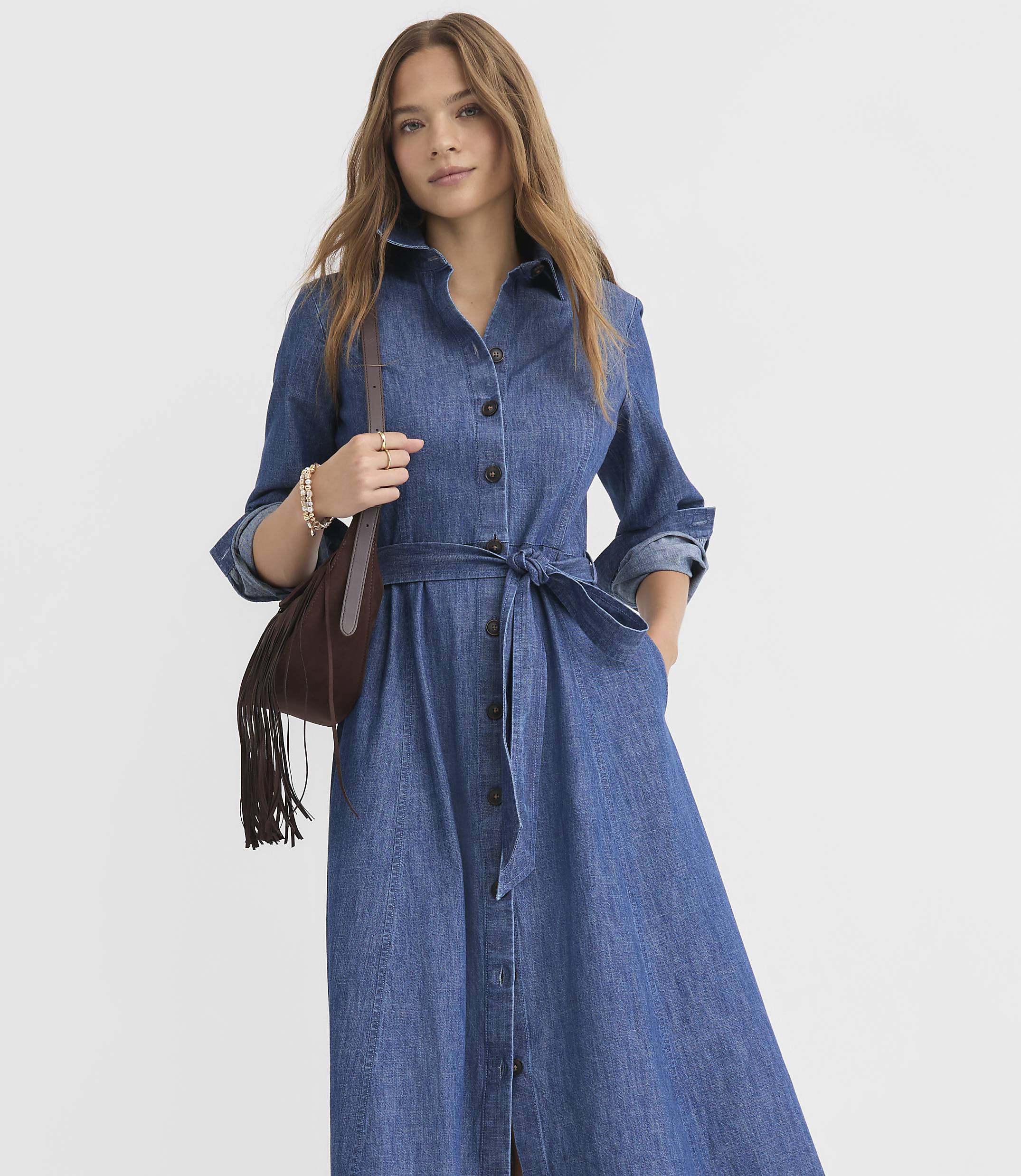 A young woman in a denim shirt dress with a fringed bag stands against a plain white background.