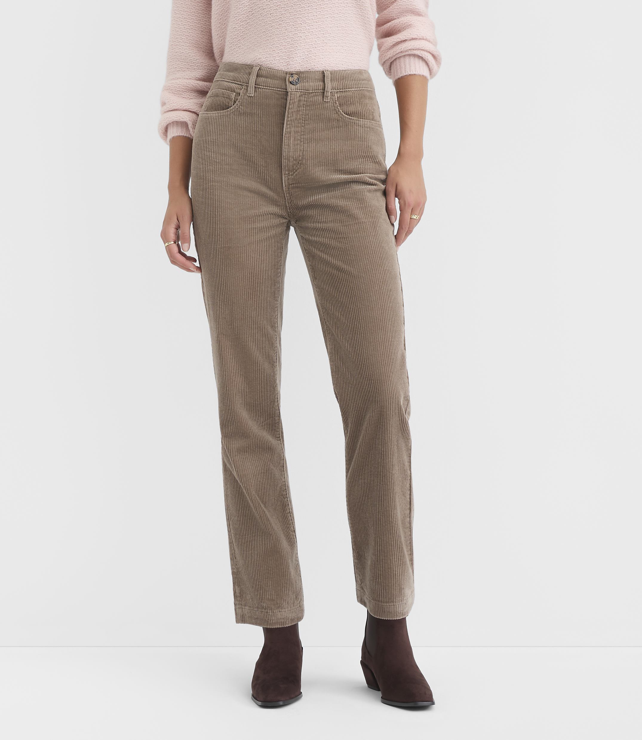 A person stands in the foreground wearing a pink sweater and tan corduroy pants, with a plain white background.