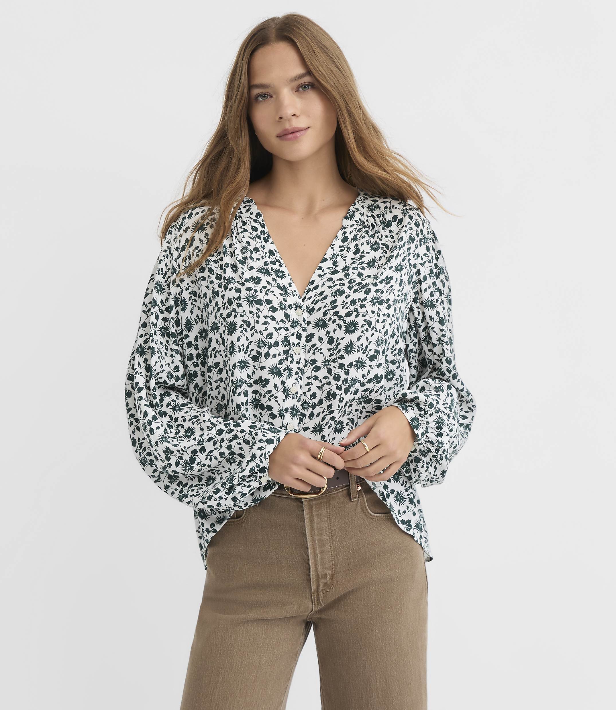 A young woman in a floral blouse and brown pants stands against a plain white background.