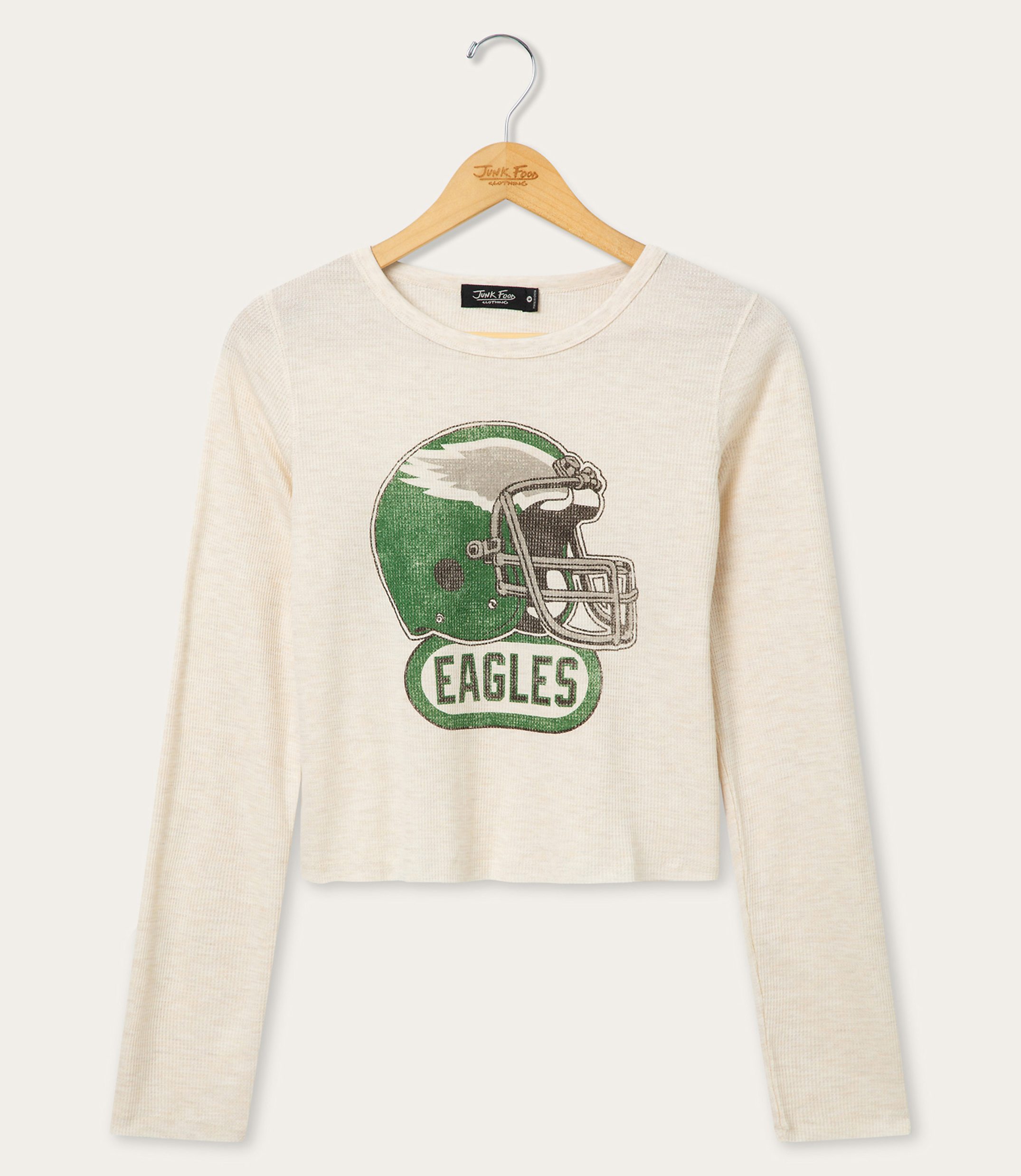 A cream-colored long-sleeved shirt with a distressed graphic of a green football helmet and the word "EAGLES" hangs on a wooden hanger against a plain white background.