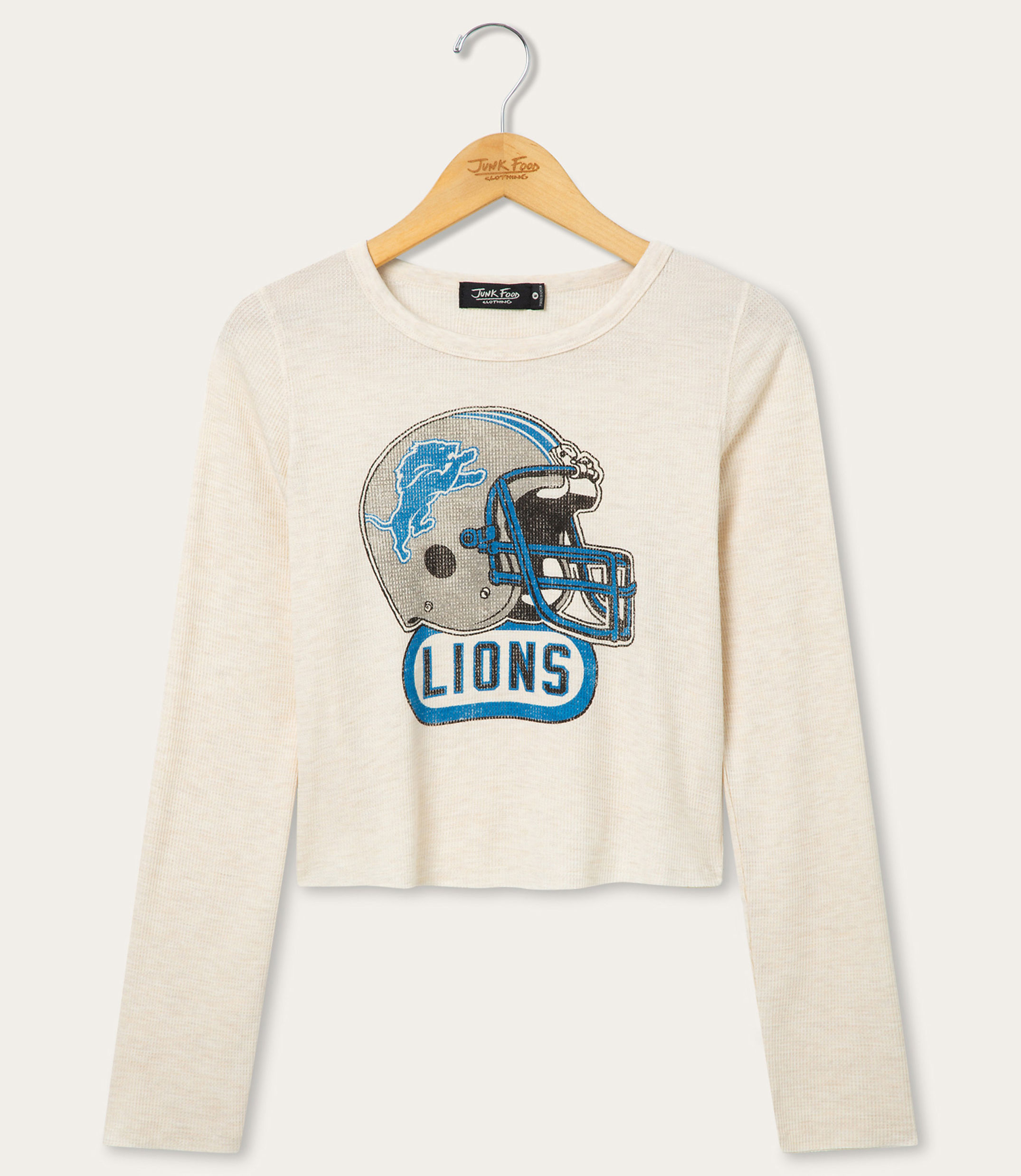 A cream-colored long-sleeved shirt with a distressed Detroit Lions helmet graphic is displayed on a hanger against a plain white background.