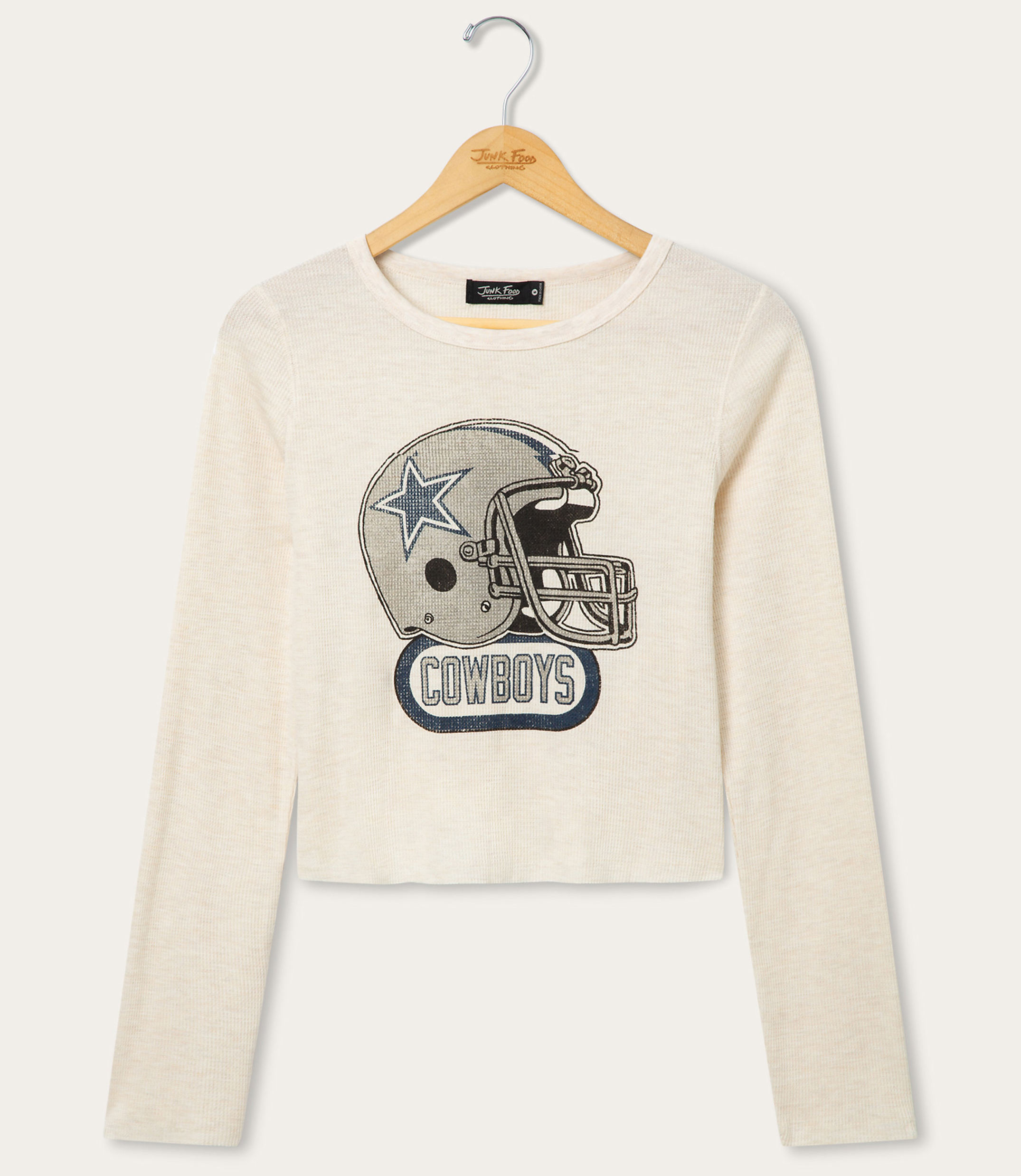 A cream-colored long-sleeved shirt with a graphic of a Dallas Cowboys helmet is displayed on a hanger against a plain white background.