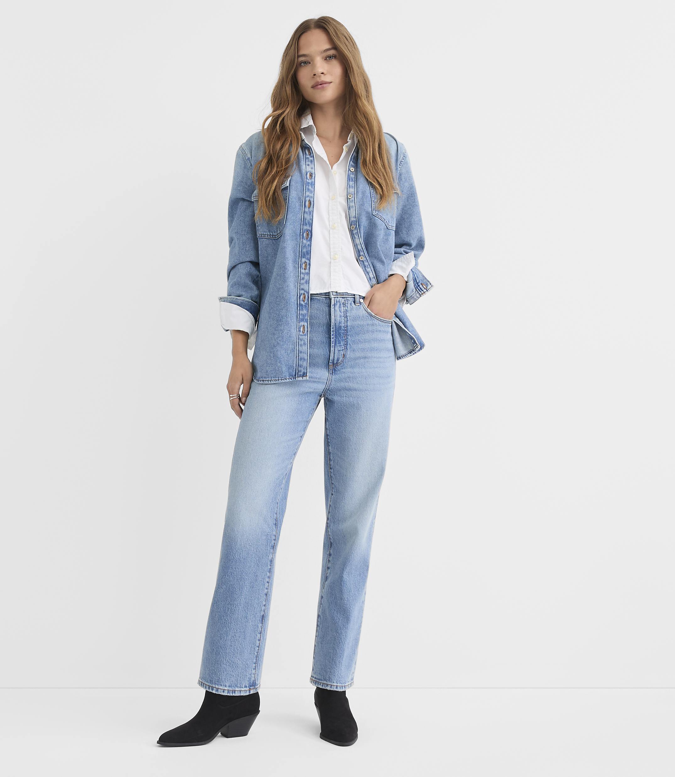 A woman in a denim shirt and jeans stands against a plain white background.