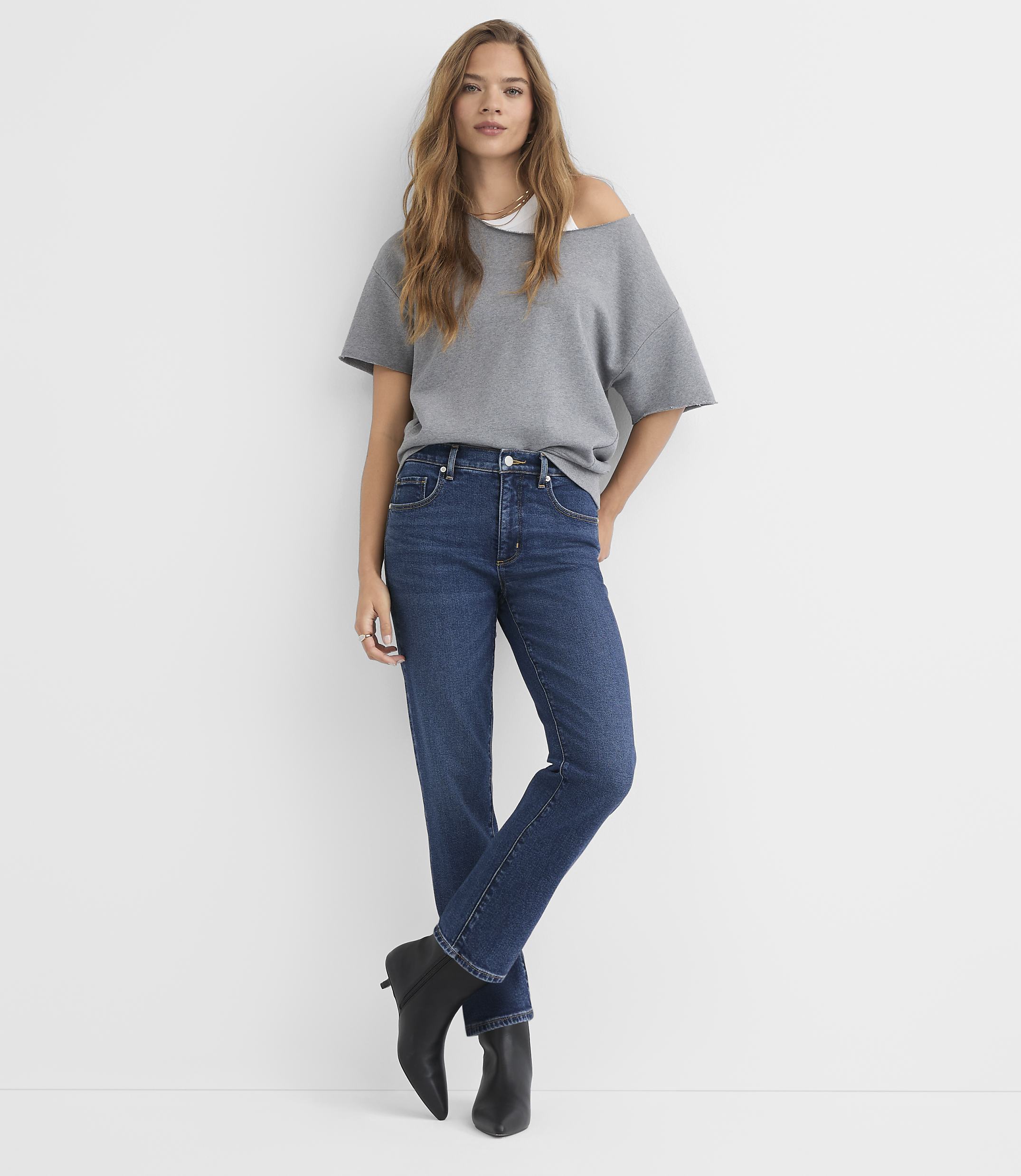 A woman in a gray off-the-shoulder top and dark wash jeans stands against a plain white background.