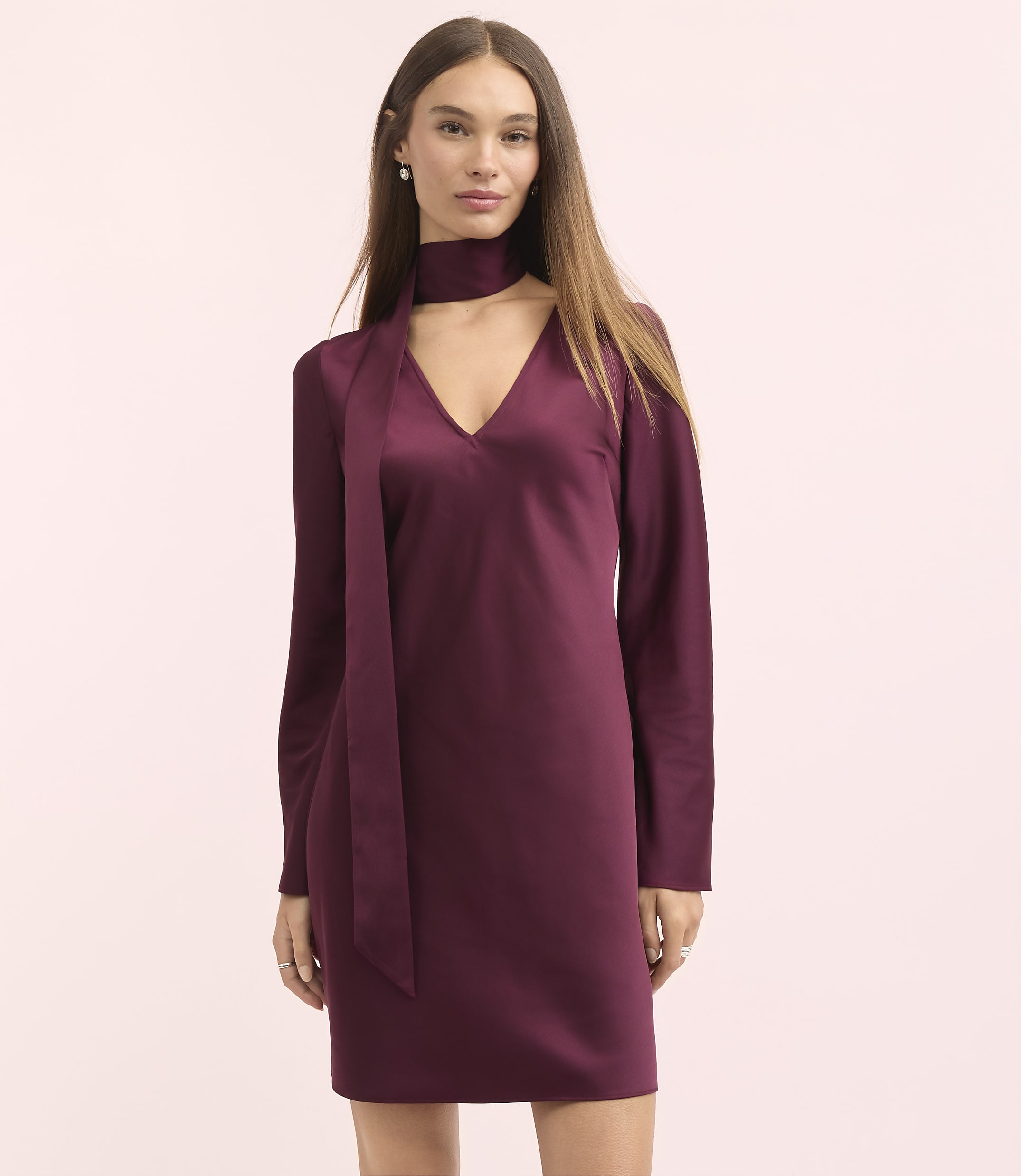 A woman in a burgundy satin dress with a choker neckline stands against a soft pink background.