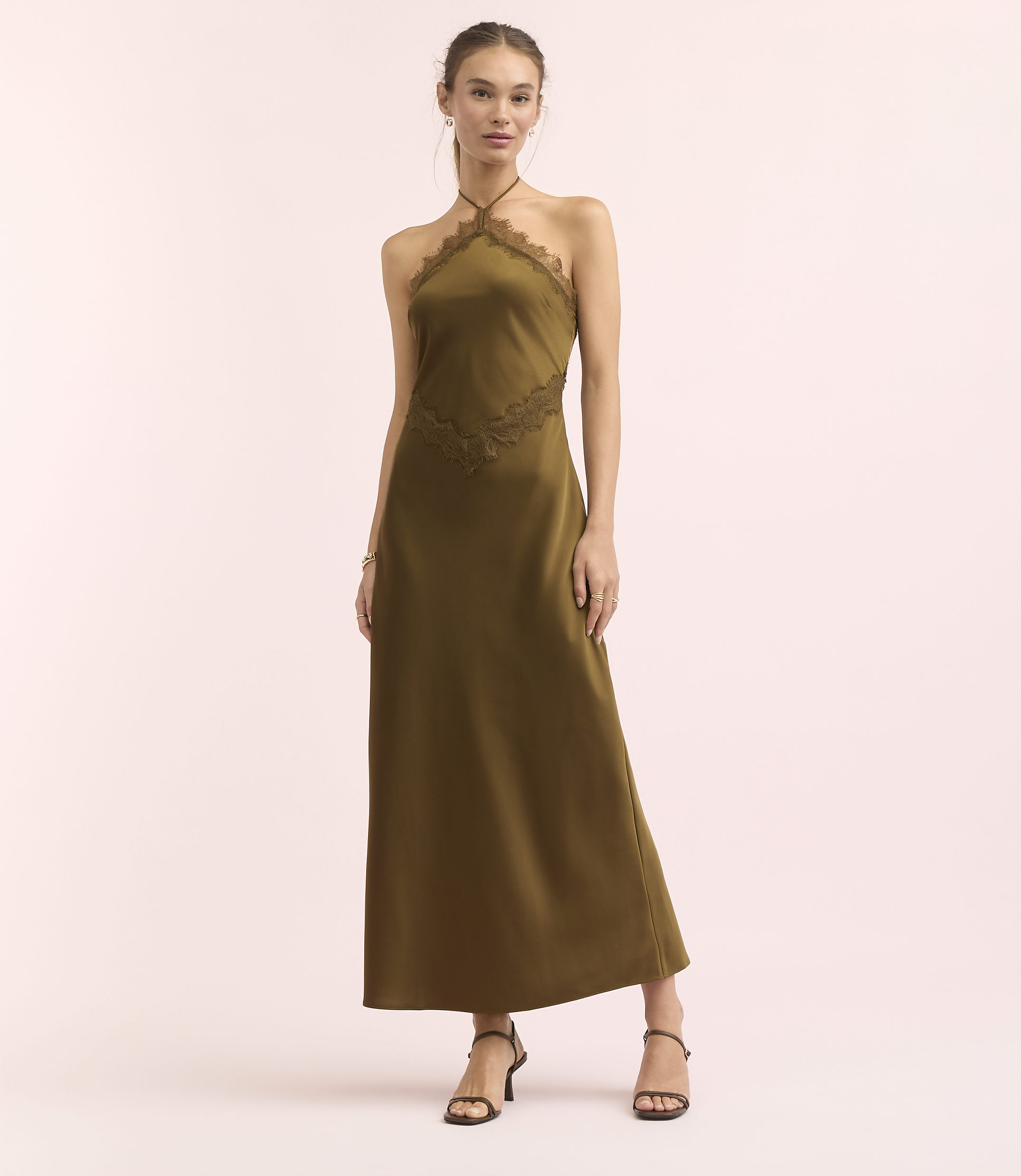 A woman in a long, olive-green halter dress with lace detailing stands against a soft pink background.