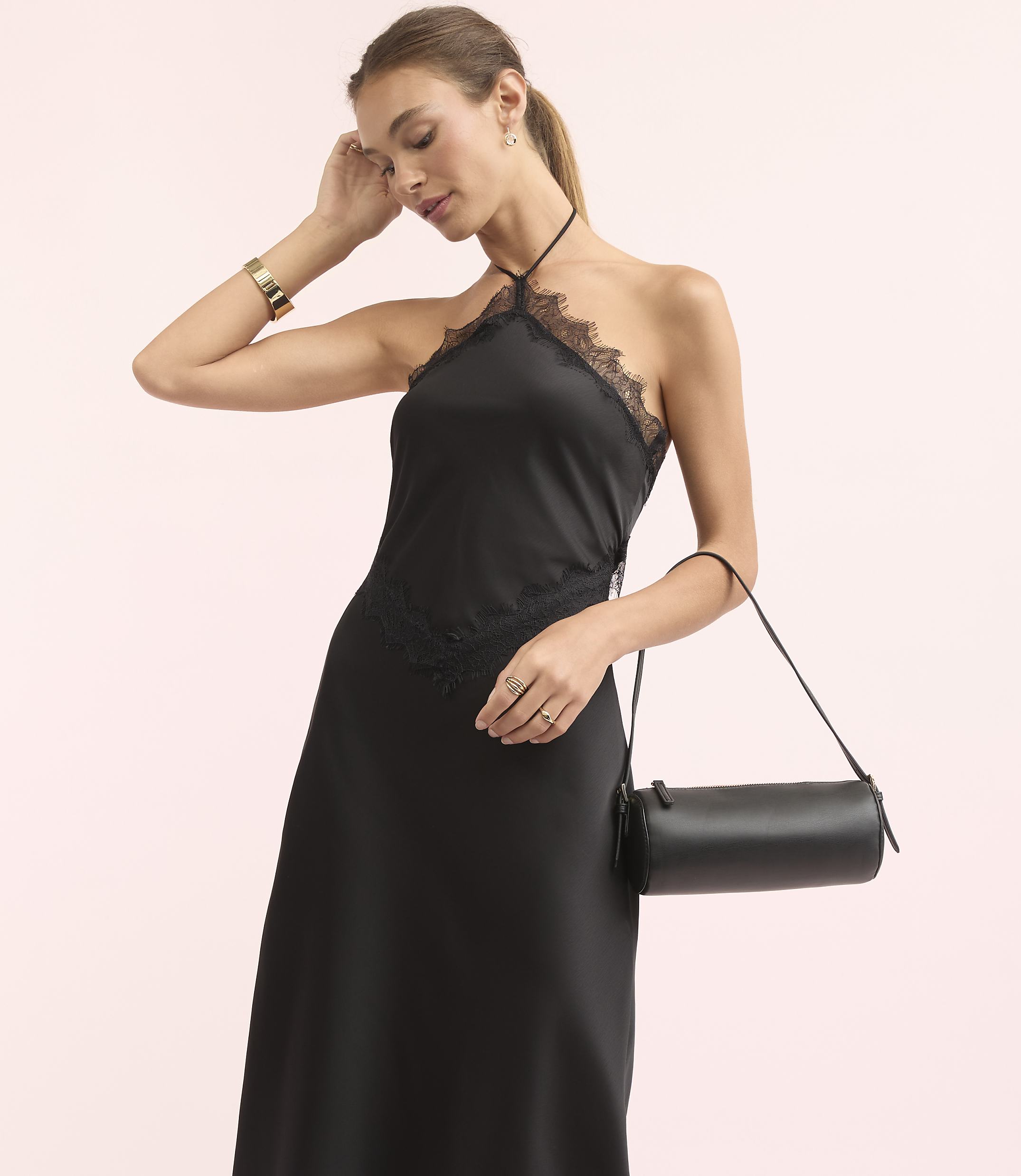 A woman in a black lace-trimmed halter dress and gold bracelet holds a cylindrical black purse against a soft pink background.