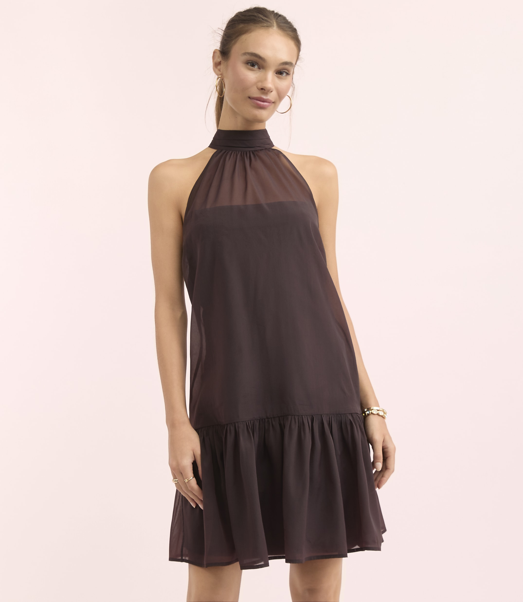 A woman in a dark brown halter dress stands against a soft pink background.