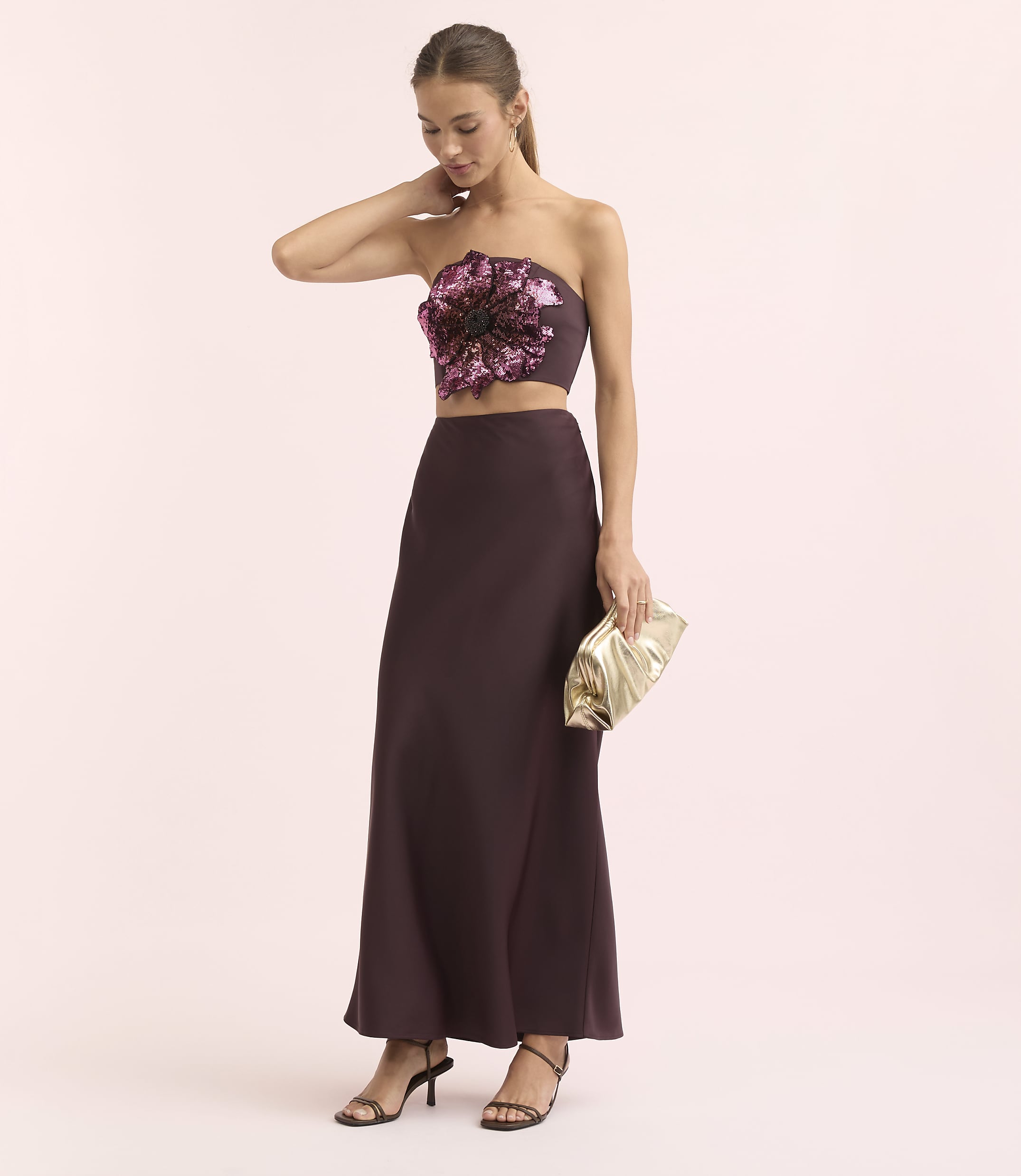 A woman in a strapless floral top and a long skirt poses with a gold clutch against a soft pink background.