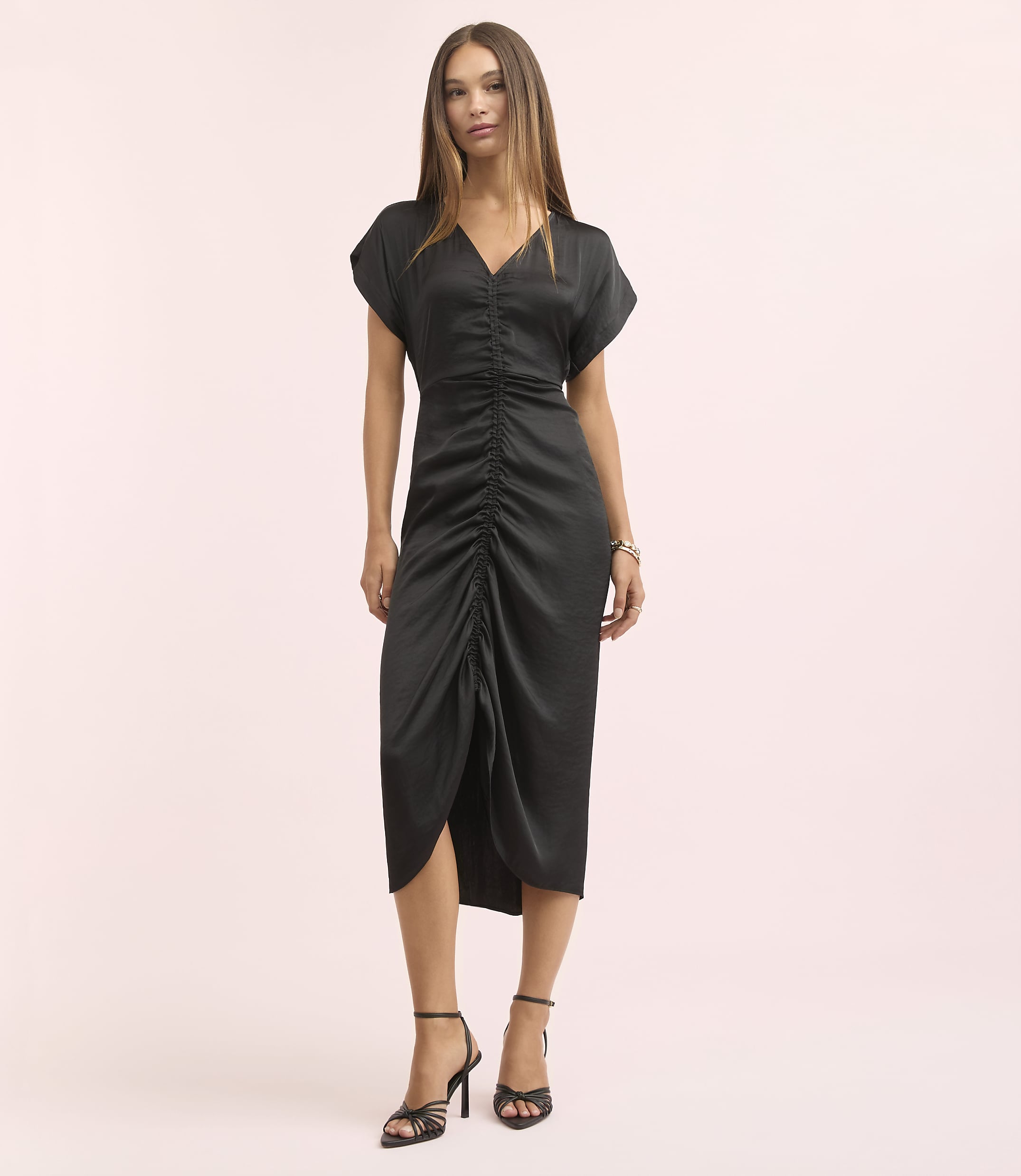 A woman in a black ruched dress and strappy heels stands against a soft pink background.