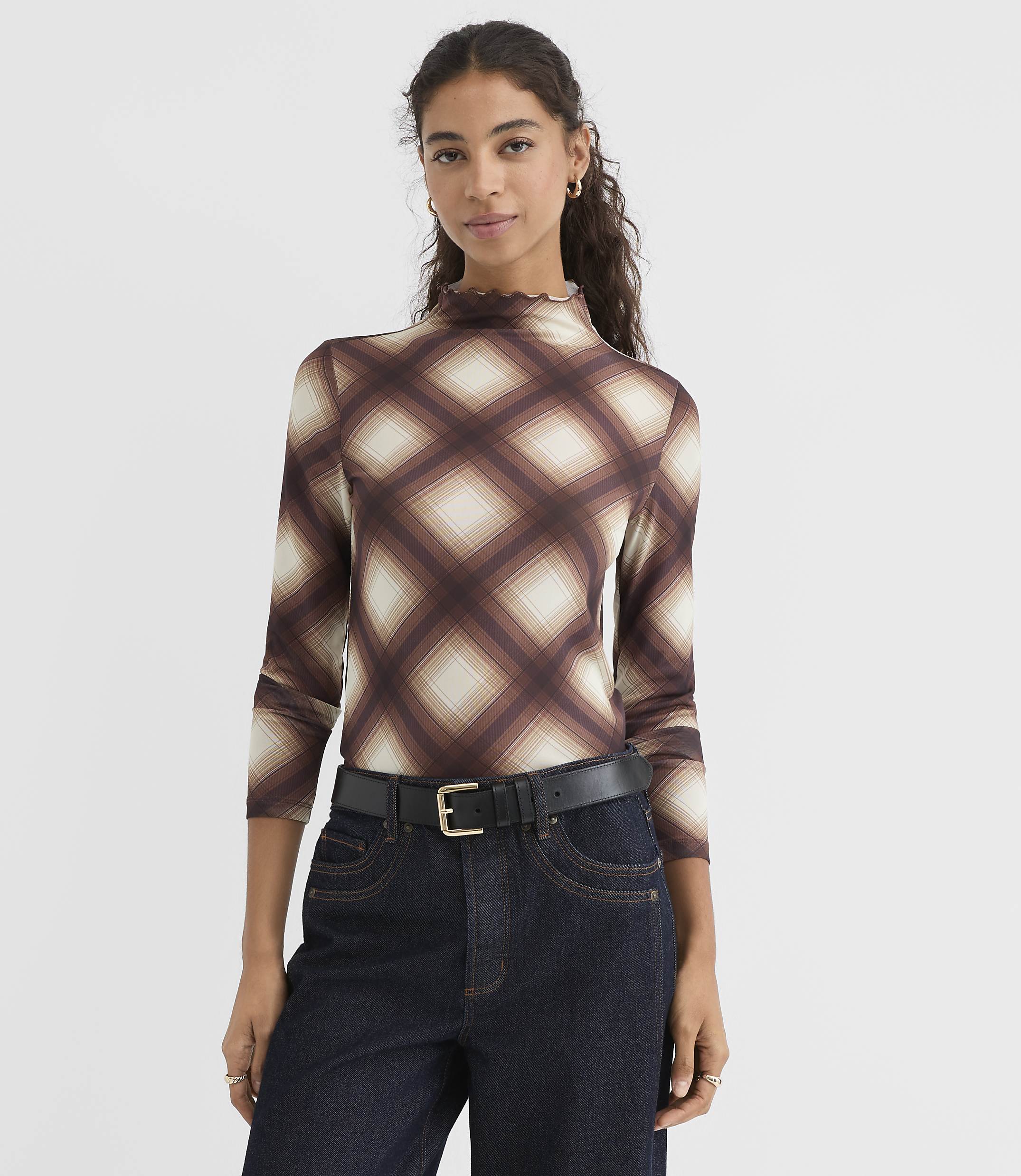 A woman in a brown plaid turtleneck and dark jeans stands against a plain white background.
