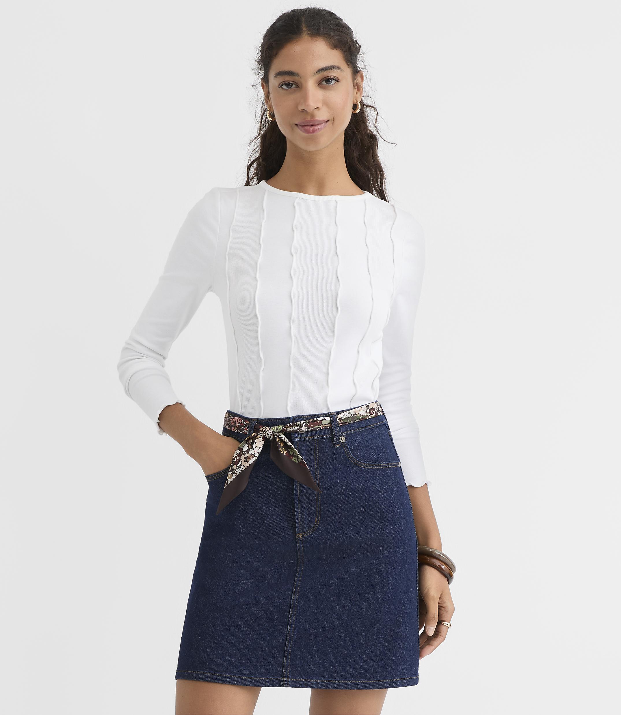 A woman in a white ribbed top and denim skirt stands against a plain white background.