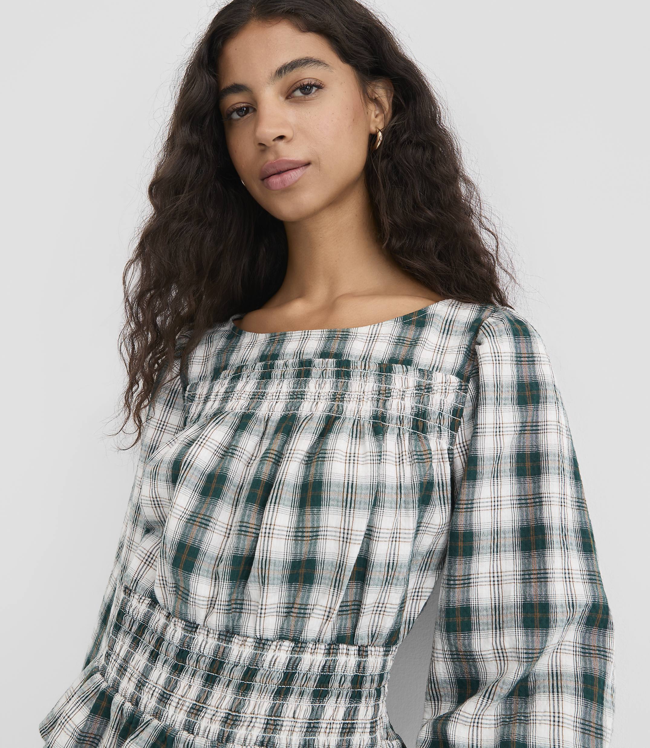 A young woman with dark curly hair wears a green and white plaid smocked top, with a plain white background.