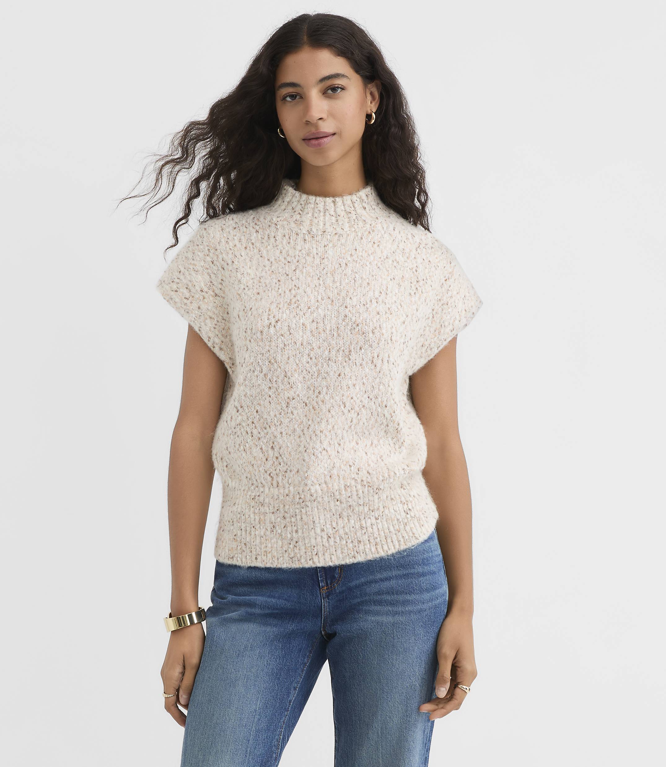 A woman in a speckled knit sweater and blue jeans stands against a plain white background.