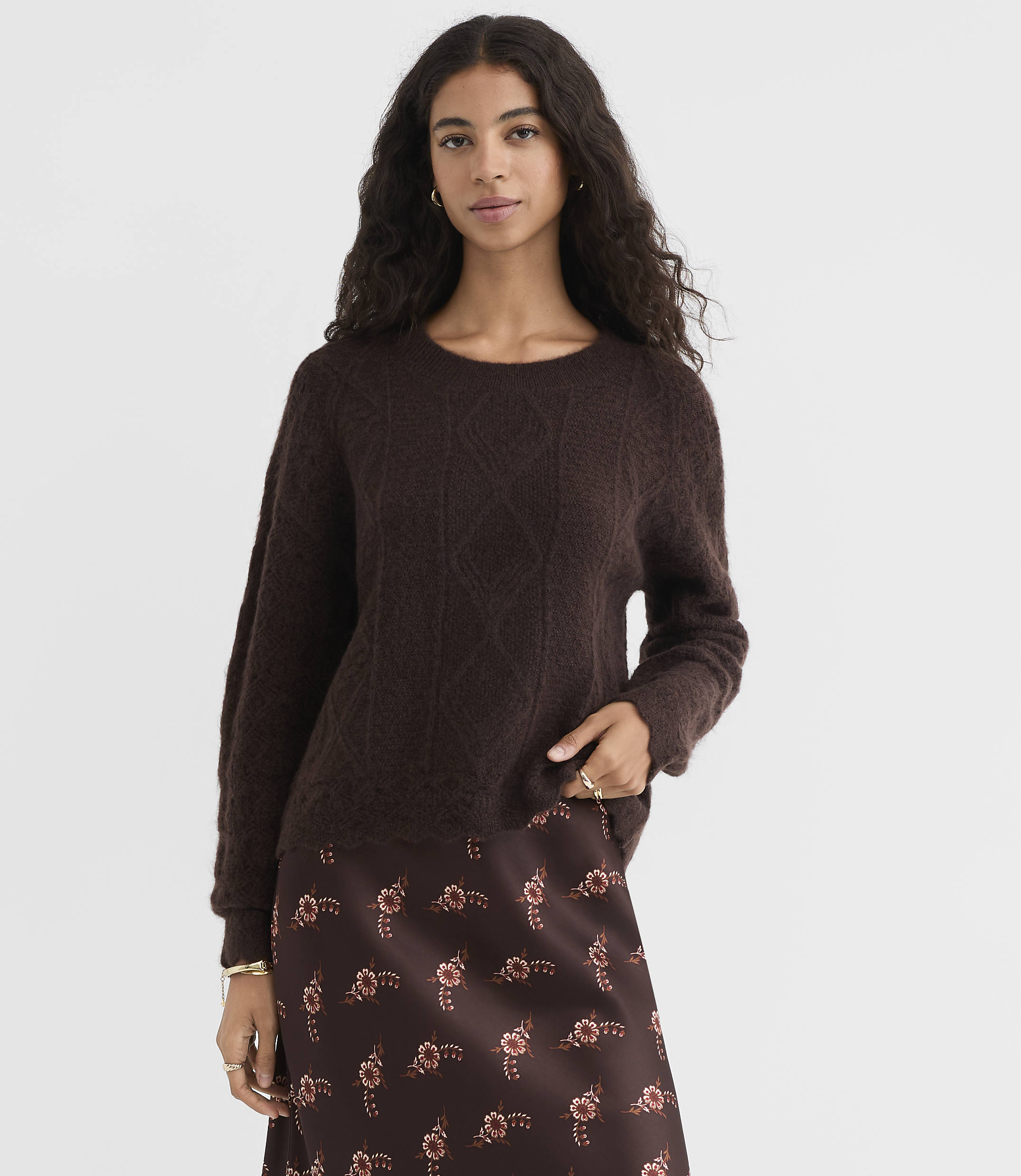 A woman in a dark brown cable-knit sweater and a floral print skirt stands against a plain, light brown background.