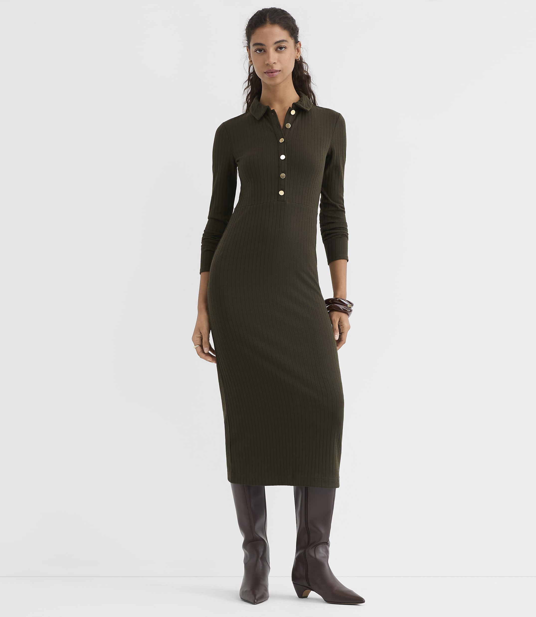A woman in a long-sleeved, olive green ribbed midi dress and brown boots stands against a plain white background.