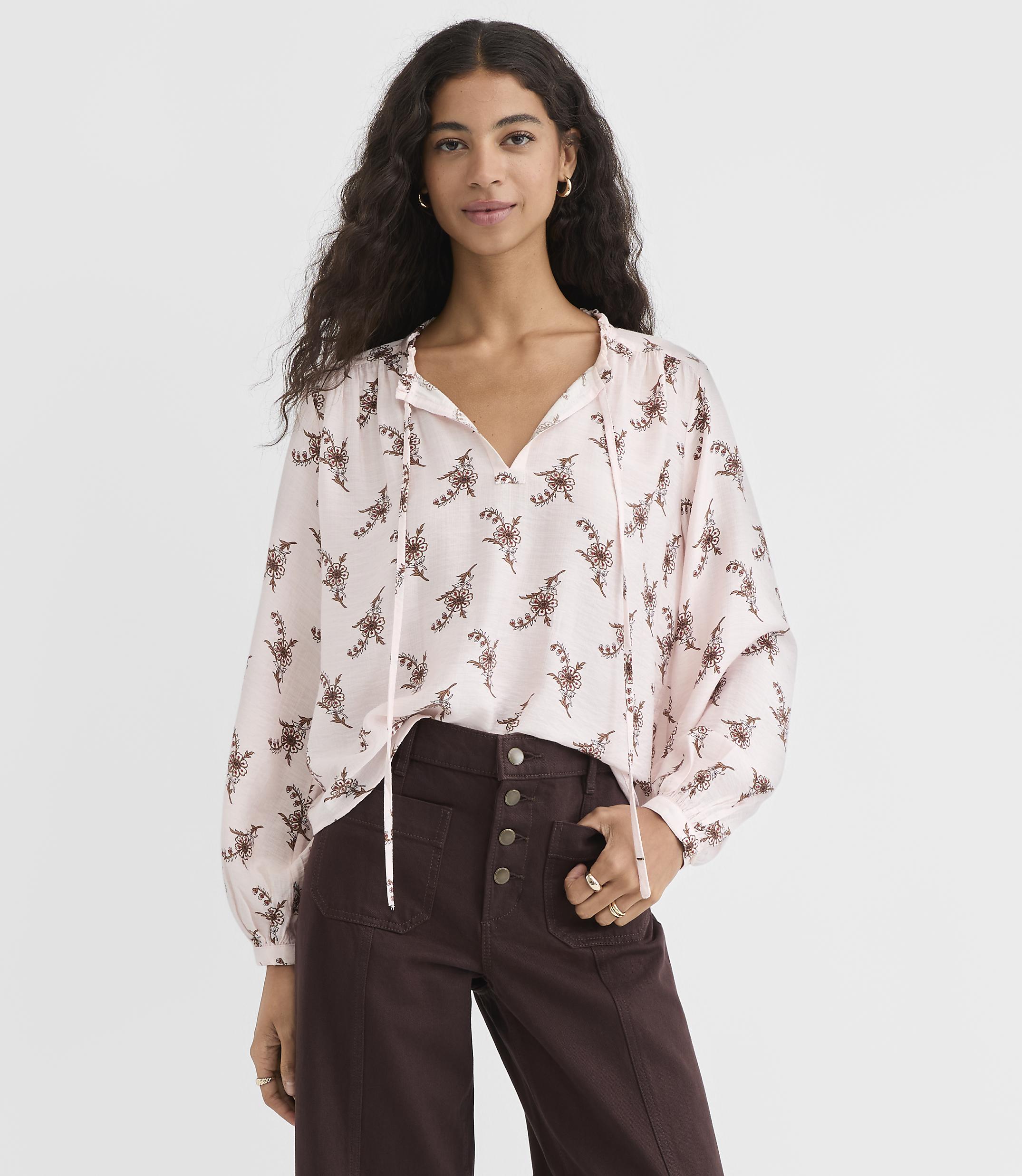A woman in a light pink, patterned blouse and dark brown pants stands against a plain white background.