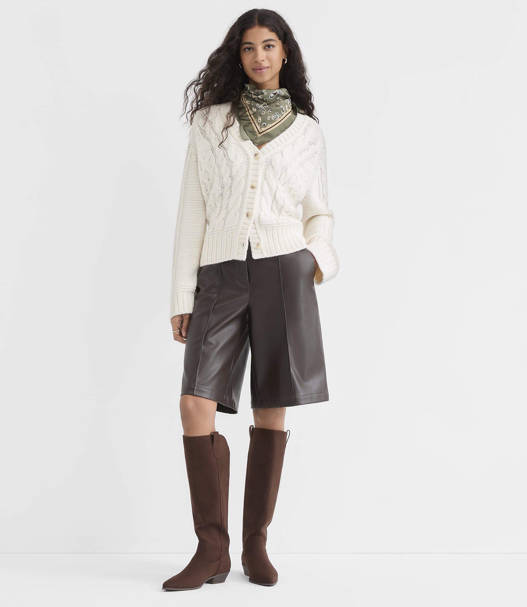 A woman in a cream cable-knit cardigan, brown leather culottes, and brown boots stands against a plain white background.