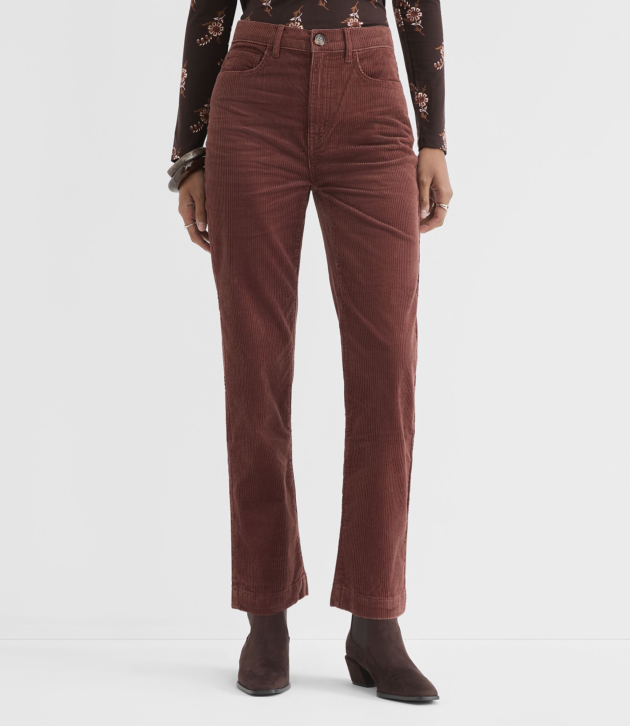 A person stands in rust-colored corduroy pants and a floral-patterned top against a plain white background.