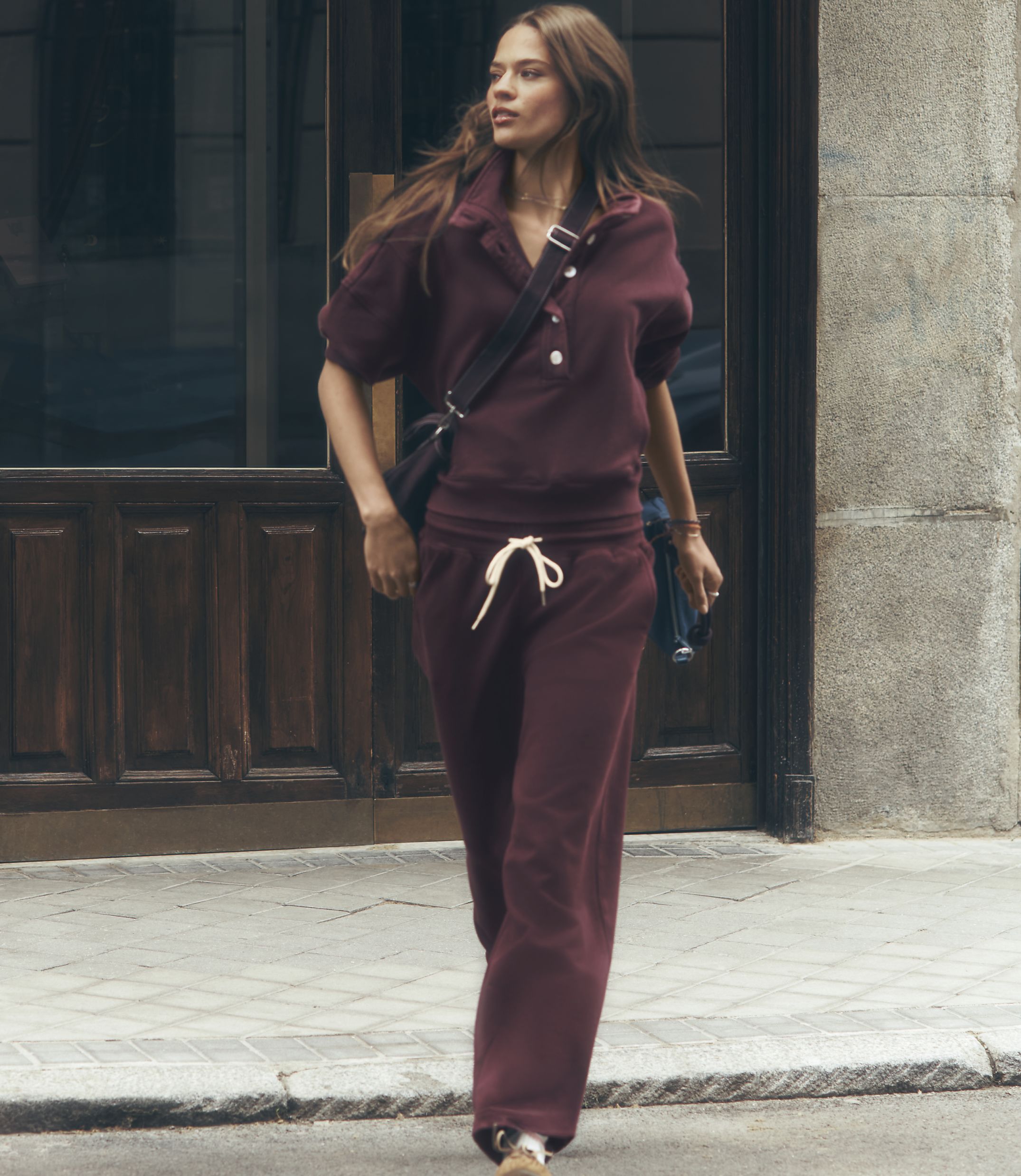 A woman in a burgundy tracksuit walks past a wooden door on a cobblestone street.