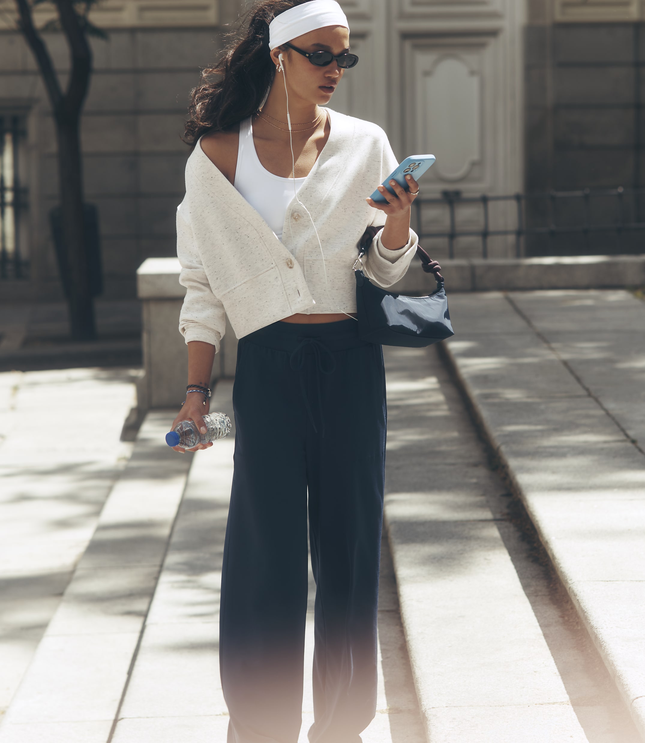 A young woman in athletic wear walks down a sunlit street, holding a water bottle and phone, with a building in the blurred background.