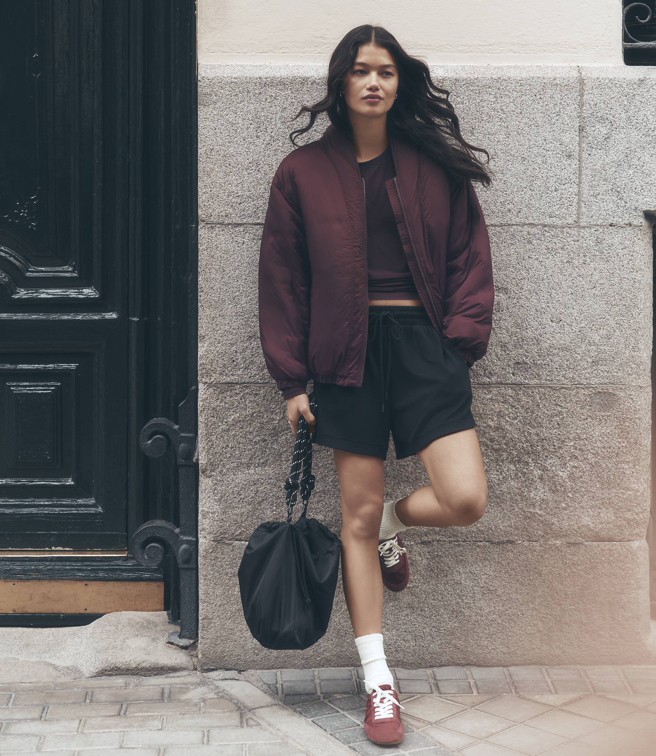 A young woman in athletic wear stands against a textured stone wall with a dark, ornate door to her left.