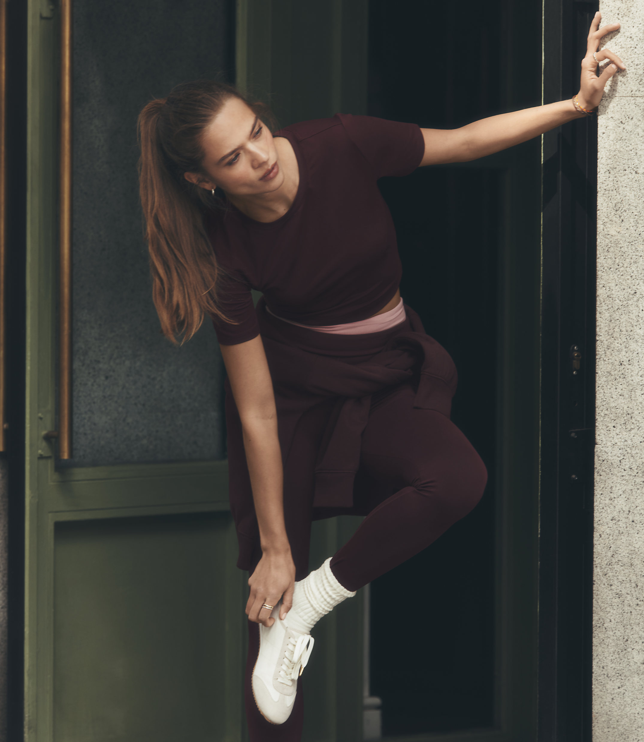 A woman in athletic wear adjusts her shoe in the foreground, with a blurred doorway and wall in the background.