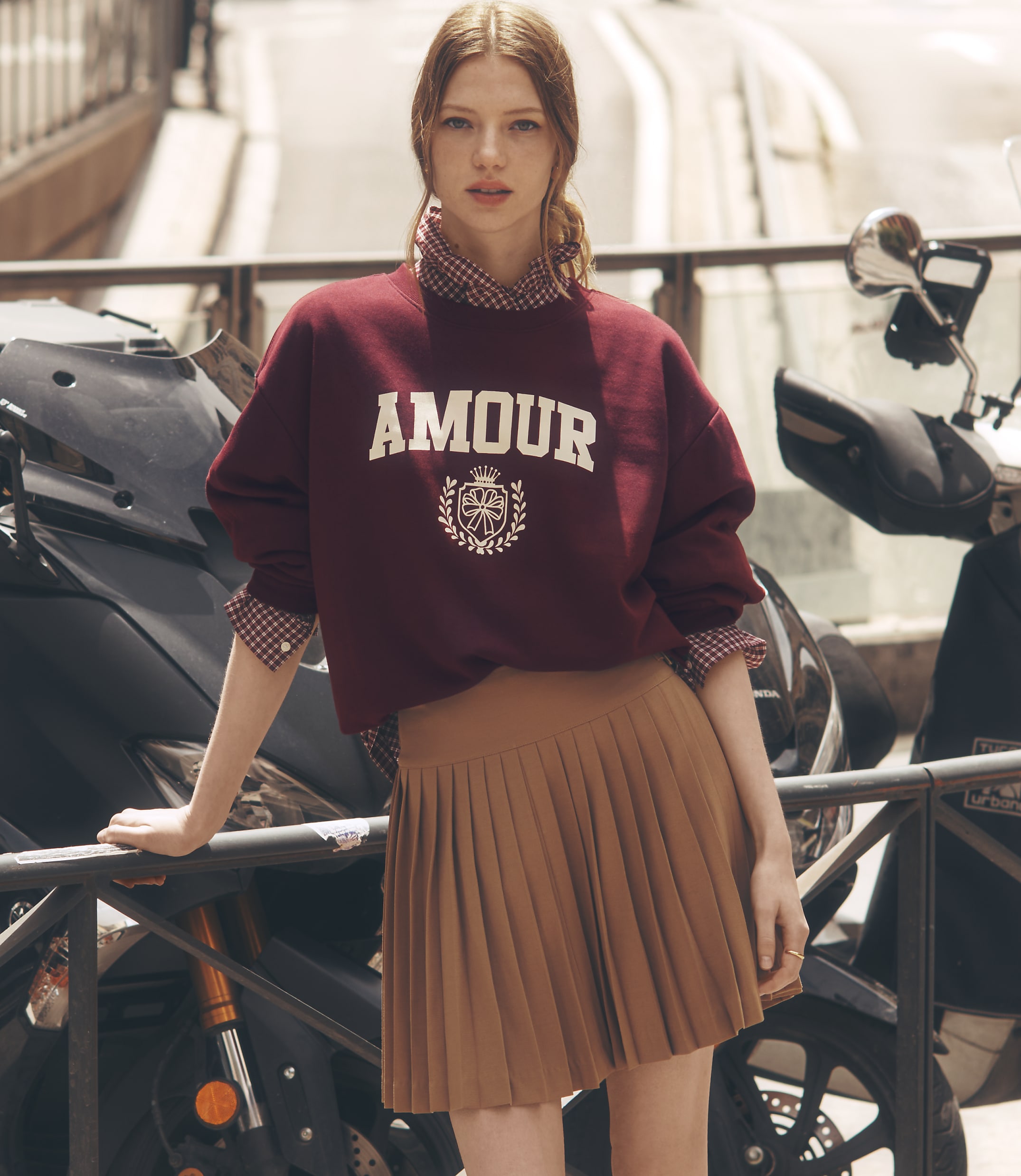 A young woman in a maroon sweatshirt and brown pleated skirt stands in front of a motorcycle with a blurred urban background.