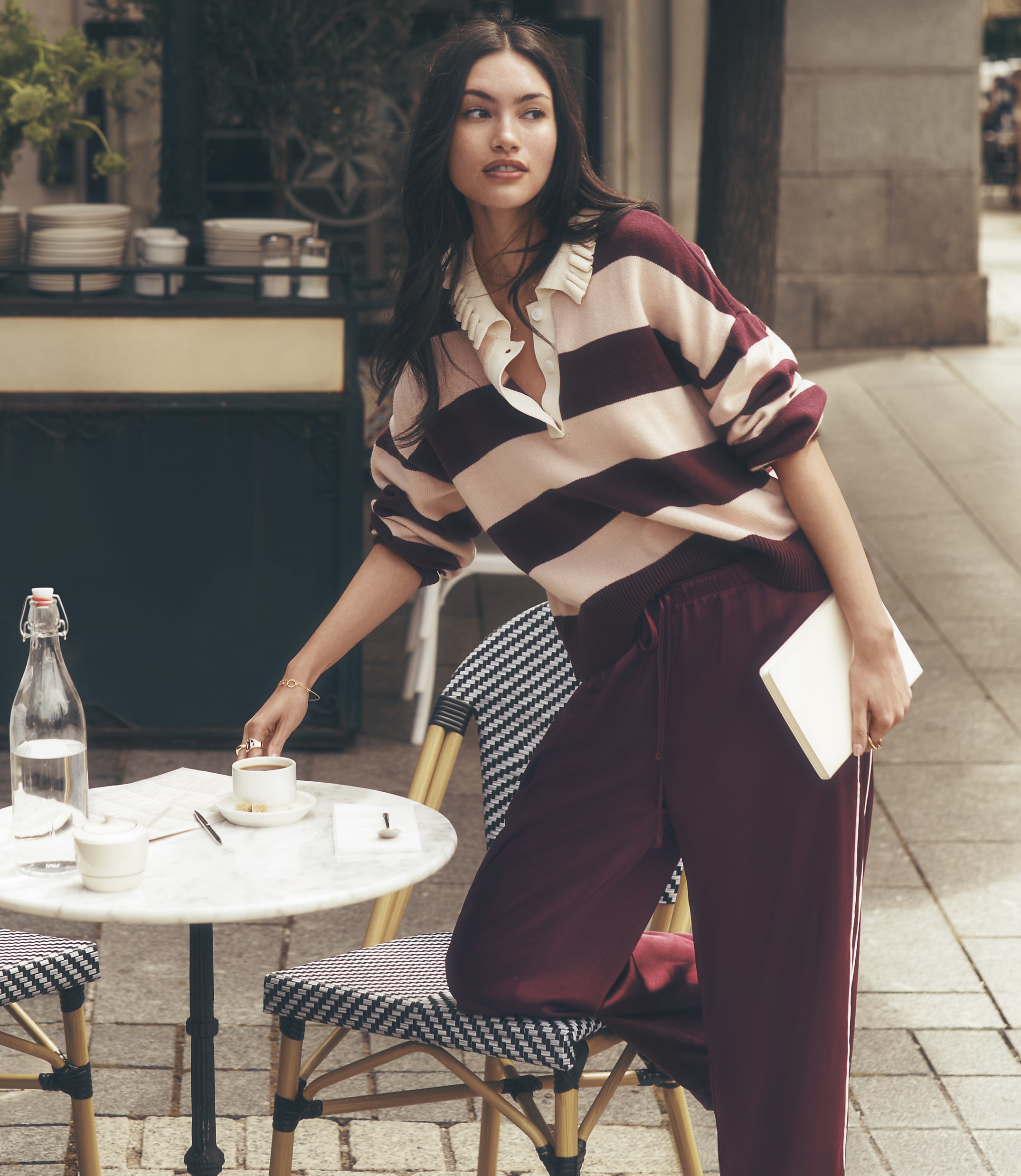A woman in a striped shirt and maroon pants leans on a cafe table with a drink and book, while a blurred street scene with buildings and a sidewalk forms the background.