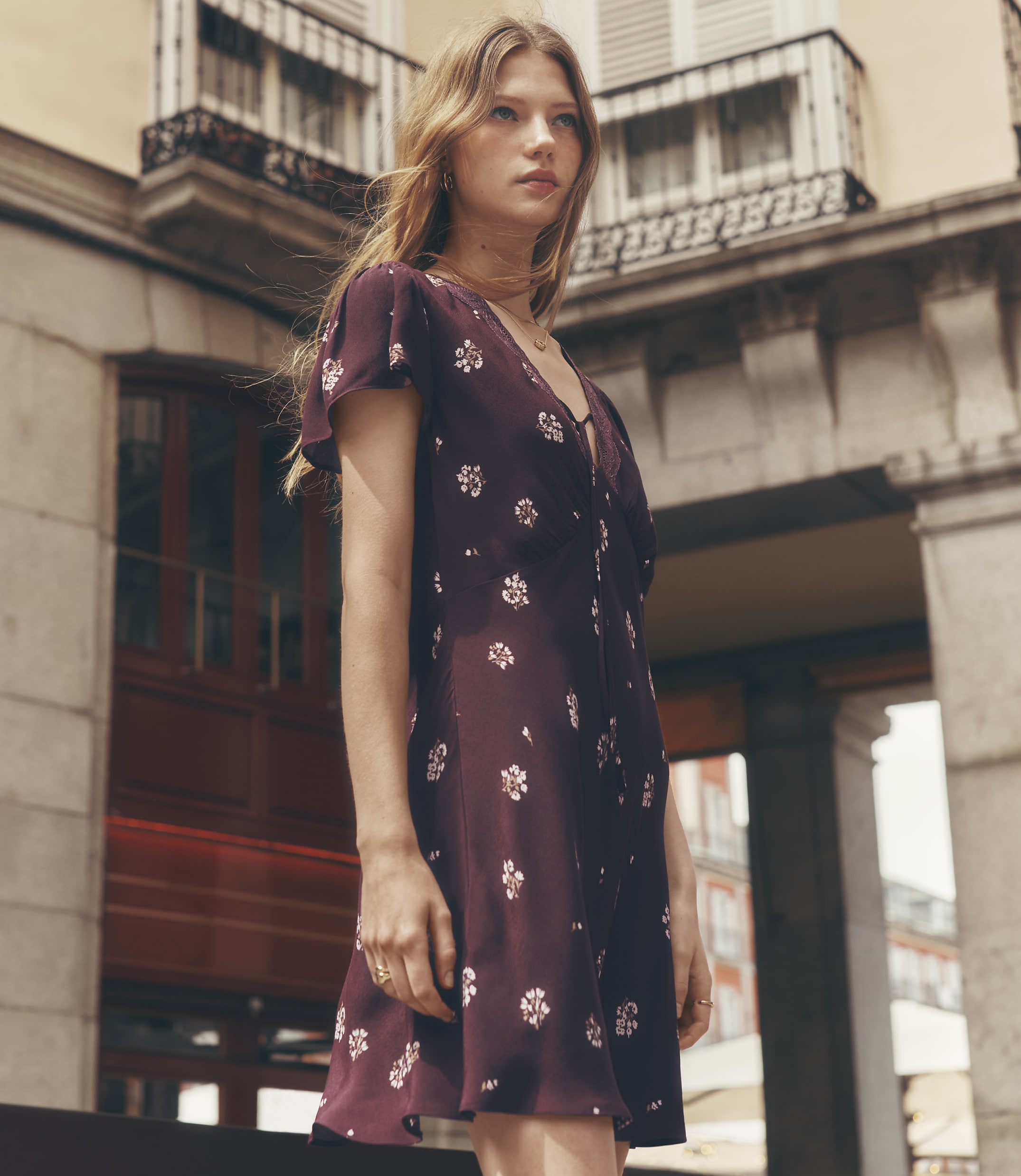 A young woman in a floral dress stands in front of a building with arched windows and balconies.