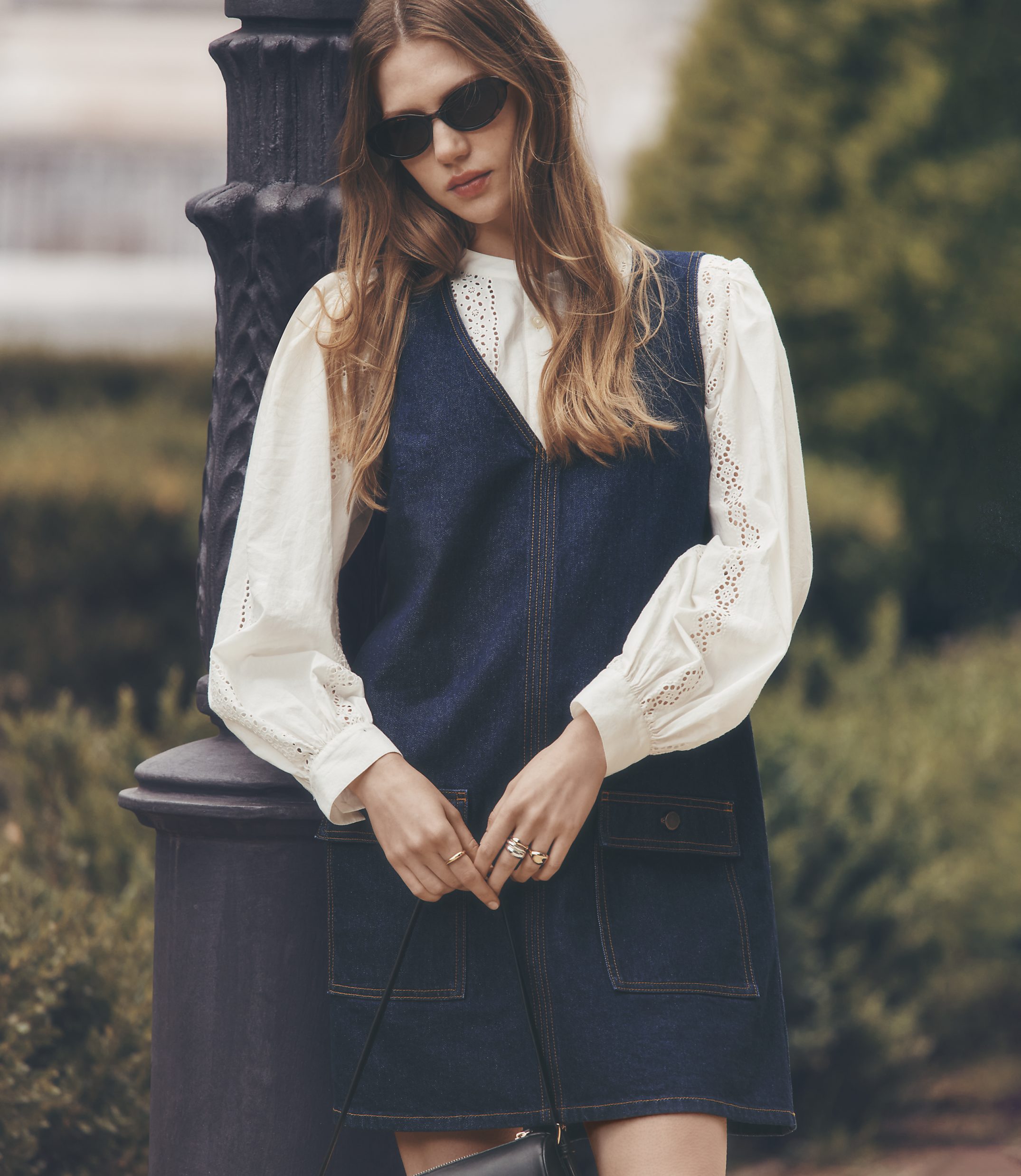 A young woman in a denim dress and white blouse leans against a lamppost with a blurred park in the background.