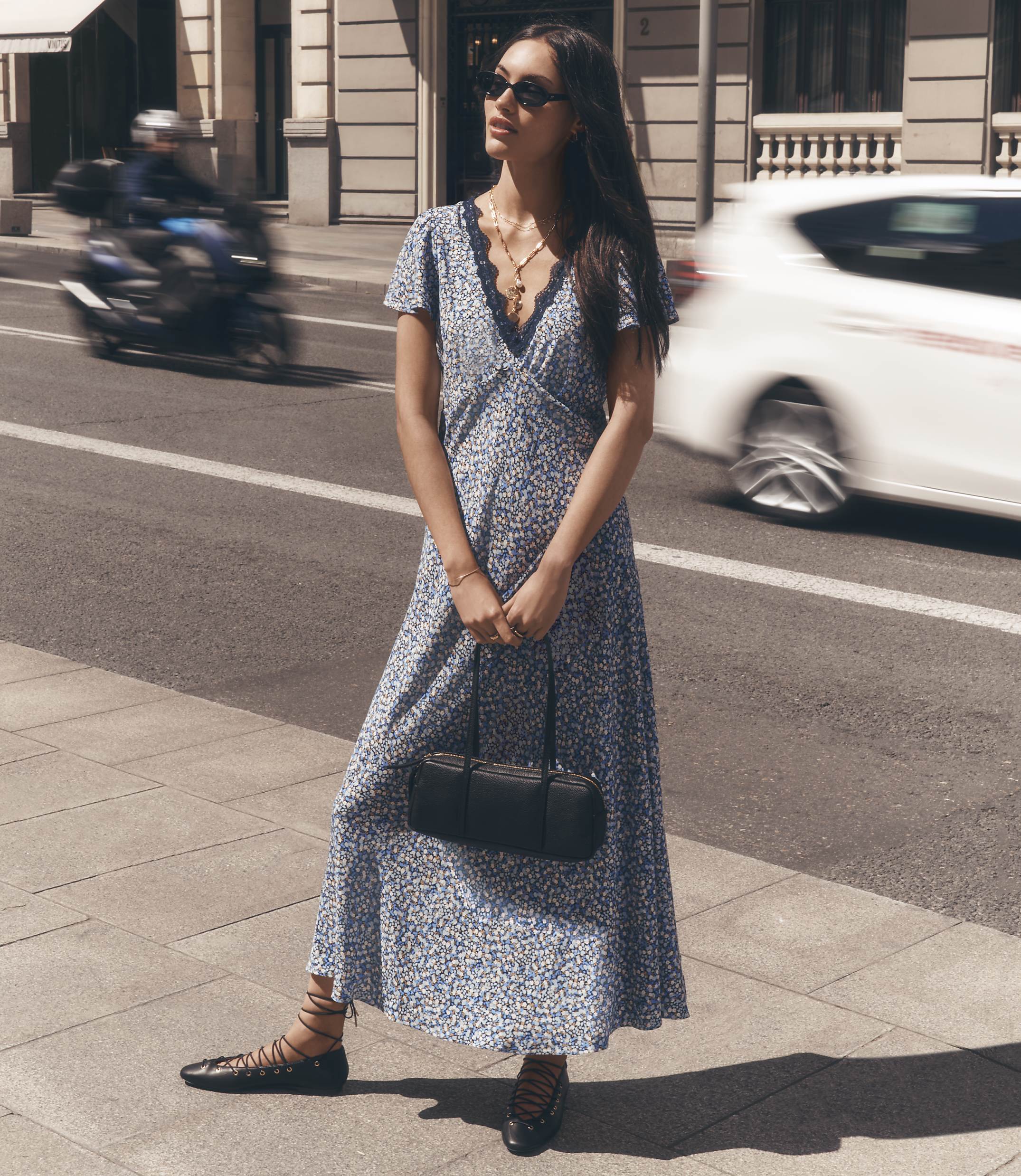 A woman in a floral dress stands on a sidewalk with a blurred motorcycle and car passing by in the background.