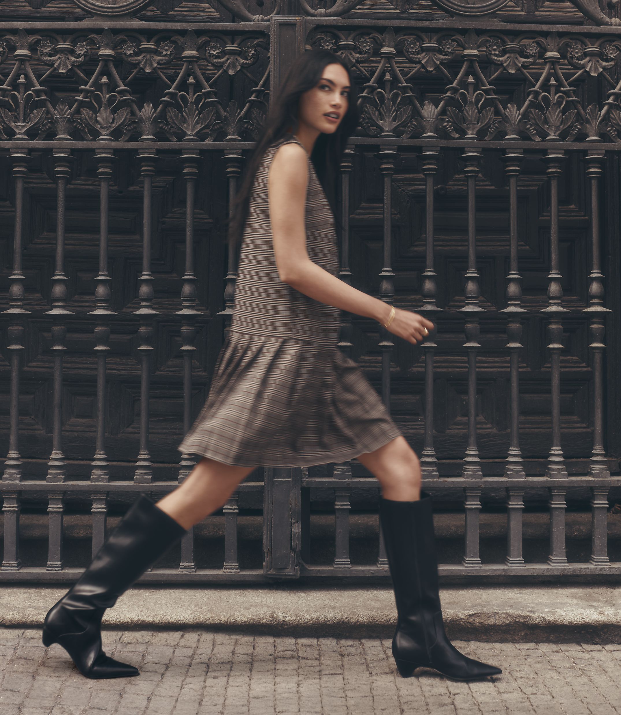 A woman in a plaid dress and black boots walks in front of an ornate metal gate.