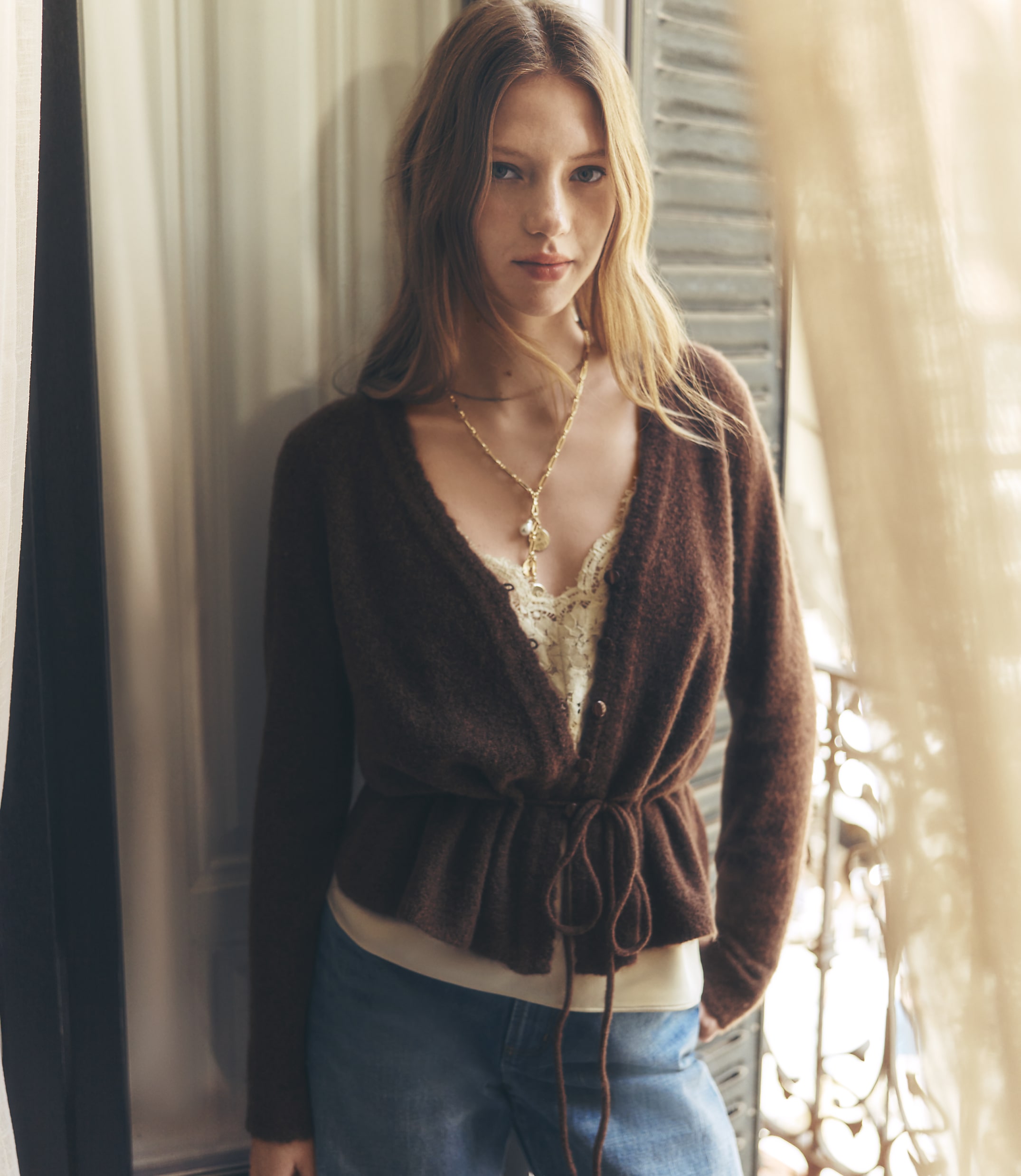 A young woman in a brown cardigan and jeans stands in front of a window with sheer curtains.