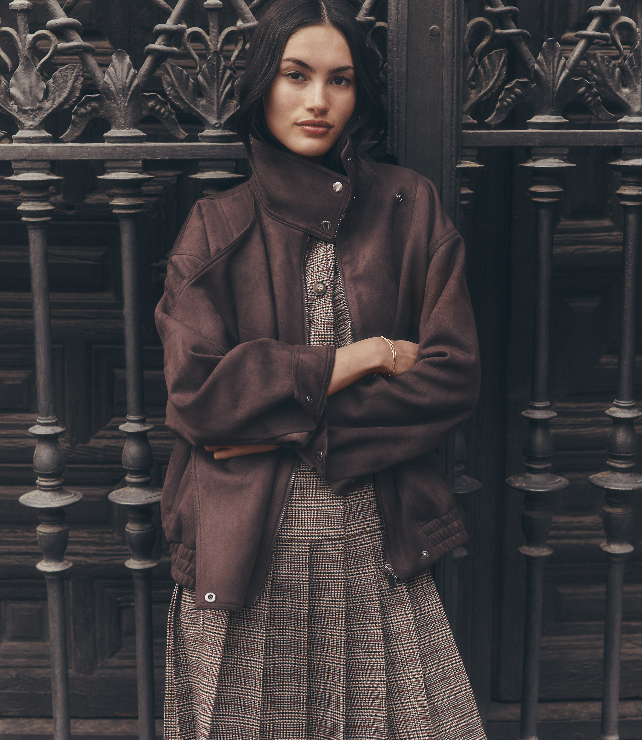 A woman in a brown jacket and plaid dress stands in front of an ornate metal gate.