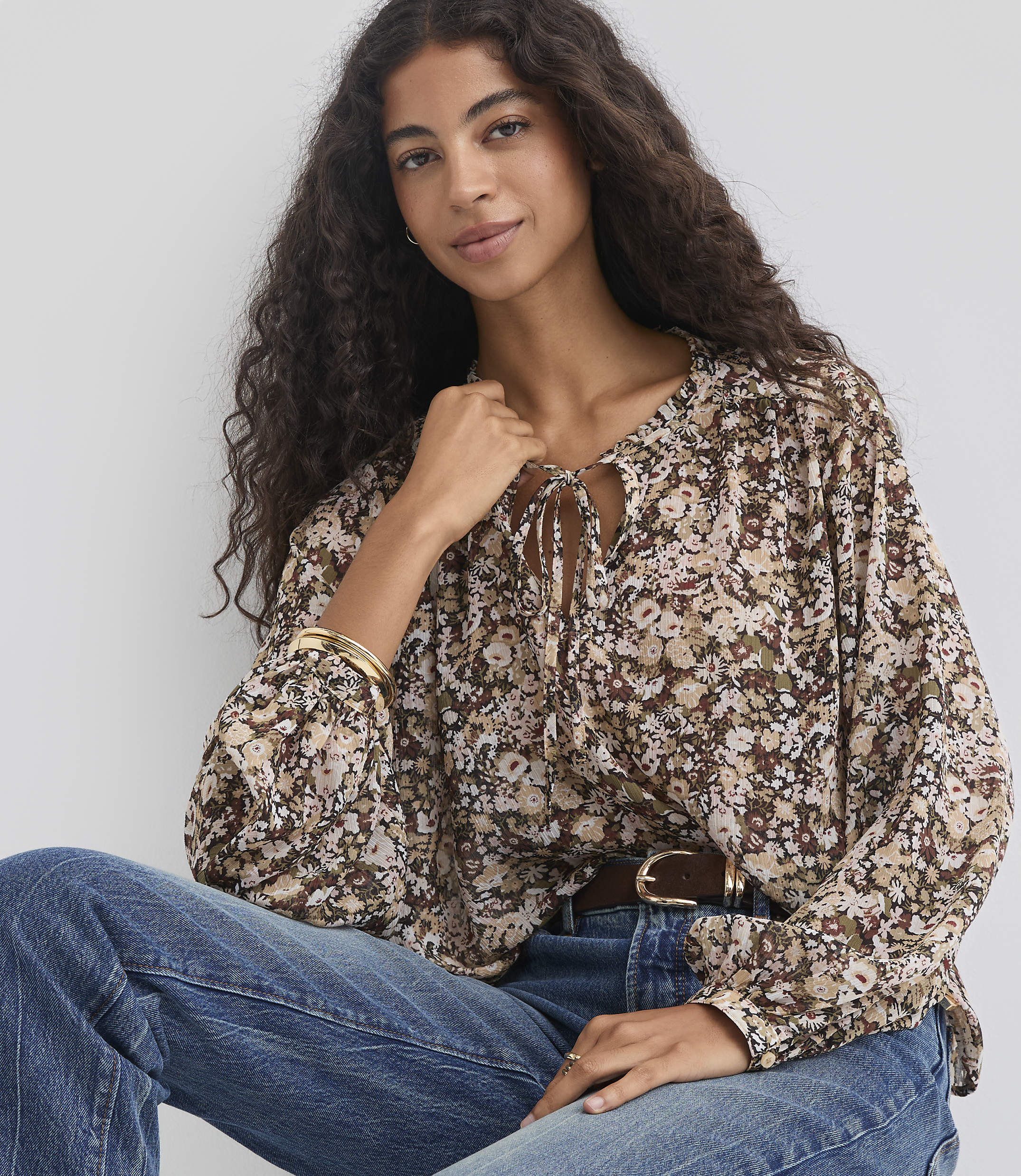 A woman with long, dark, curly hair wears a floral print blouse and blue jeans, seated against a plain, light-colored background.
