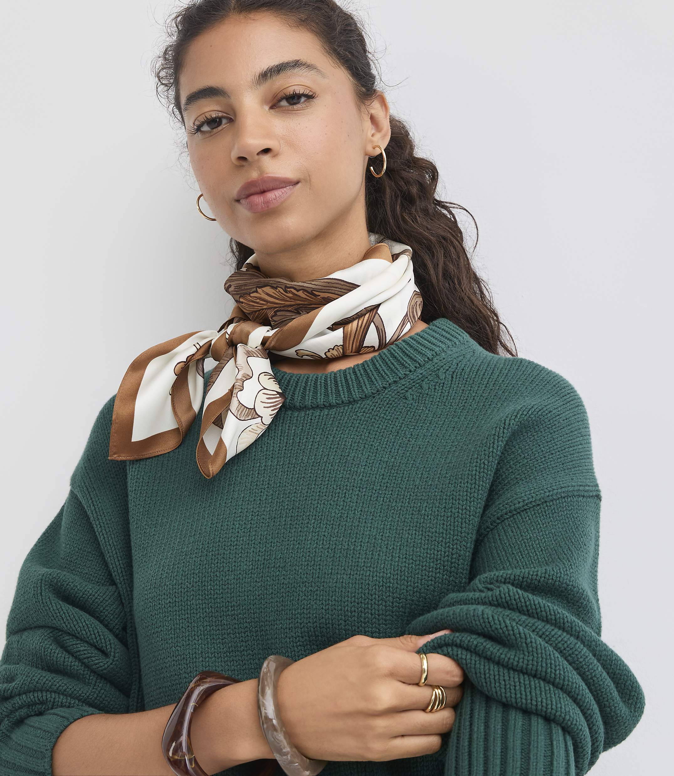 A woman with curly dark hair and a patterned silk scarf around her neck poses in front of a plain white background.