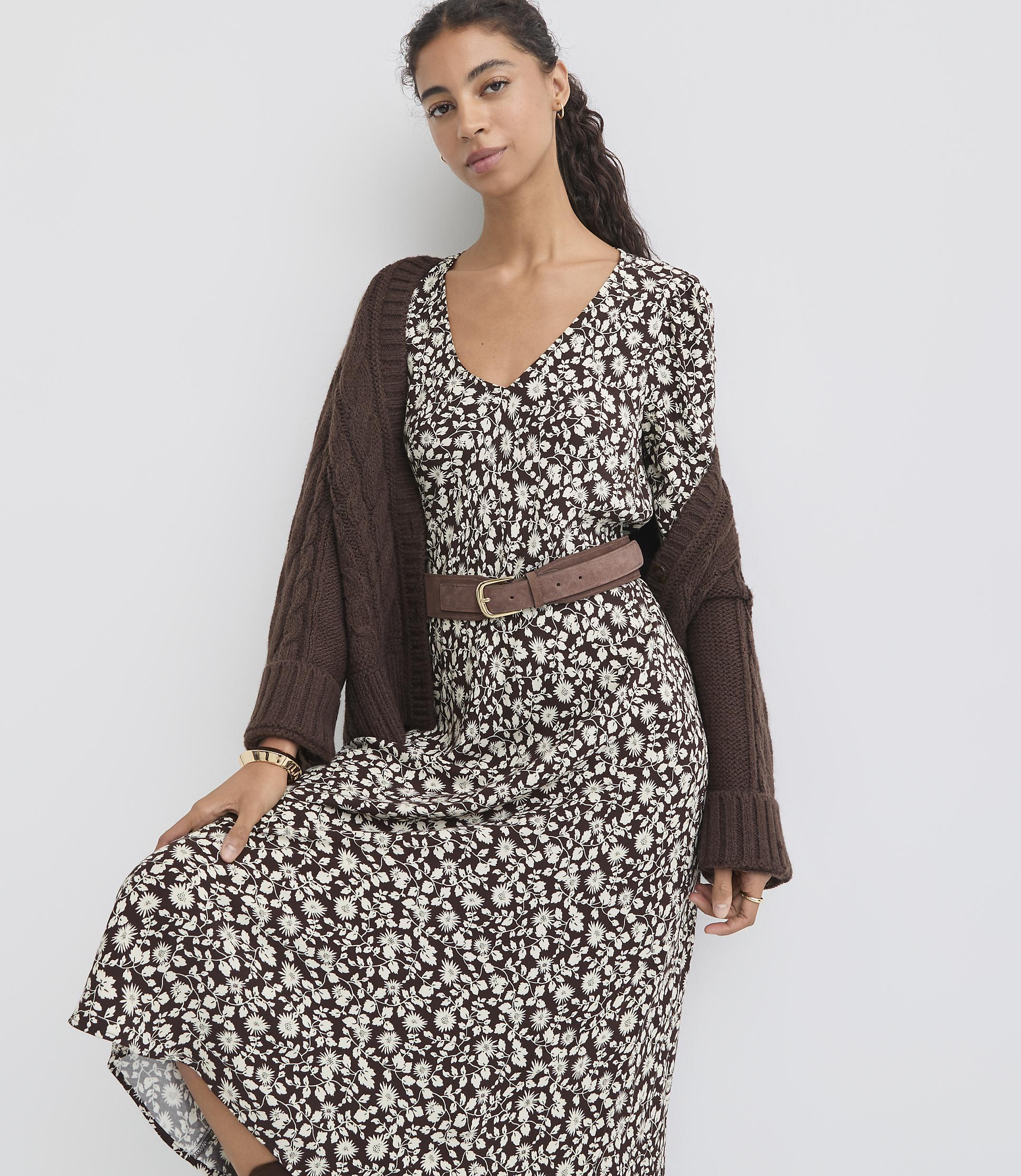 A woman in a floral dress and brown cardigan stands against a plain white background.