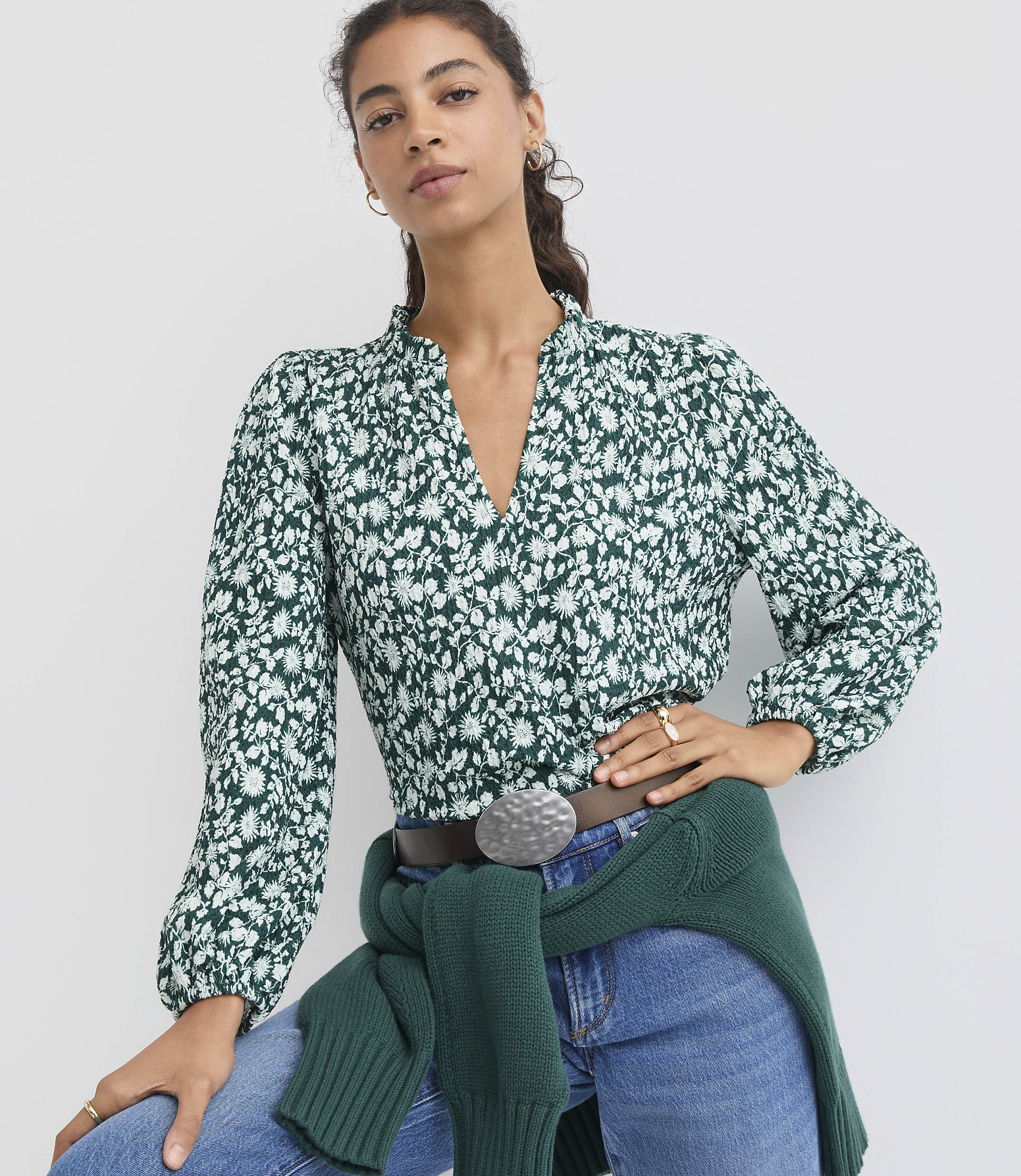 A woman in a floral blouse and jeans stands against a plain white background.