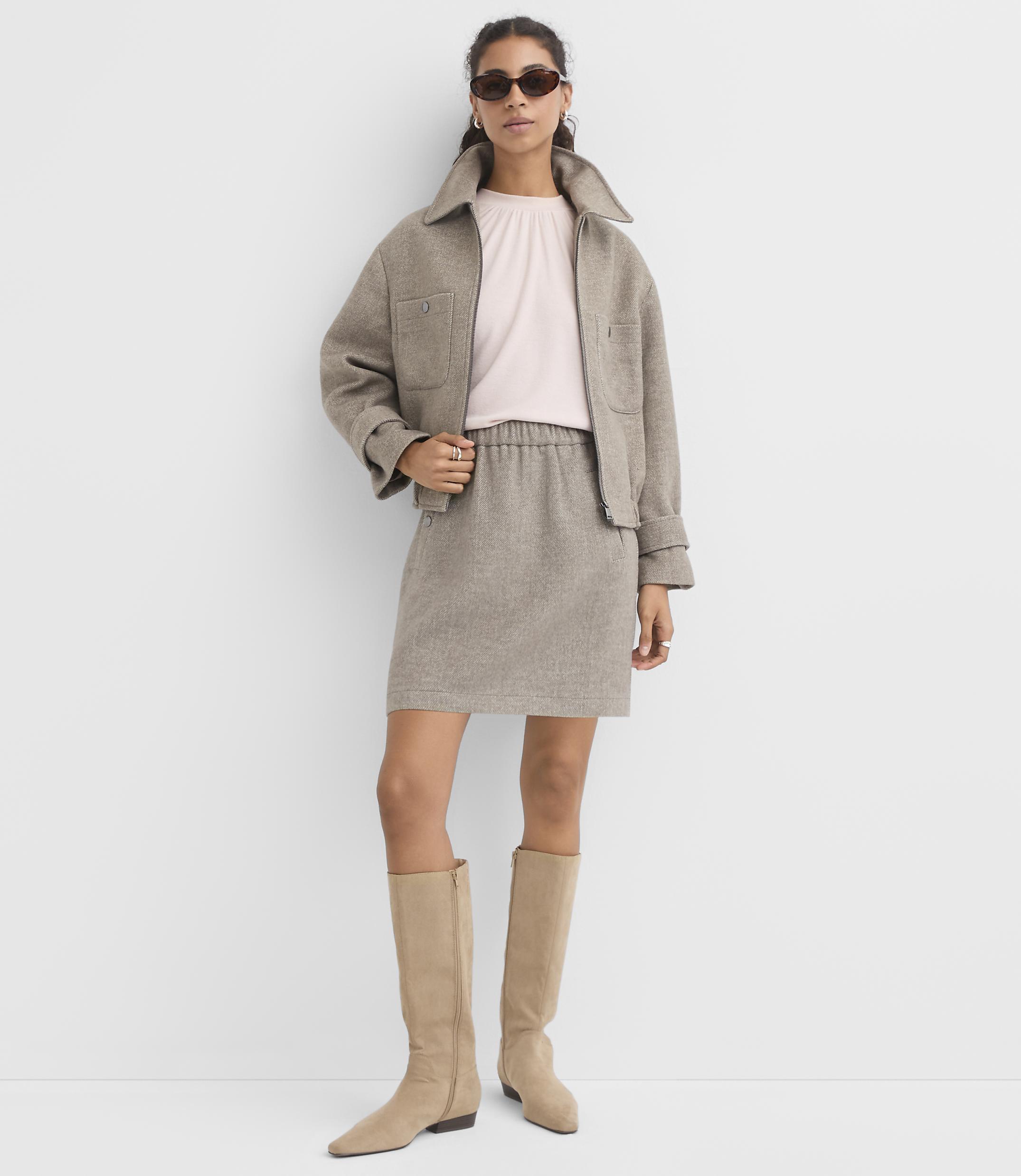 A woman in a light pink top, beige jacket, and matching skirt stands against a plain white background, wearing knee-high beige boots and sunglasses.