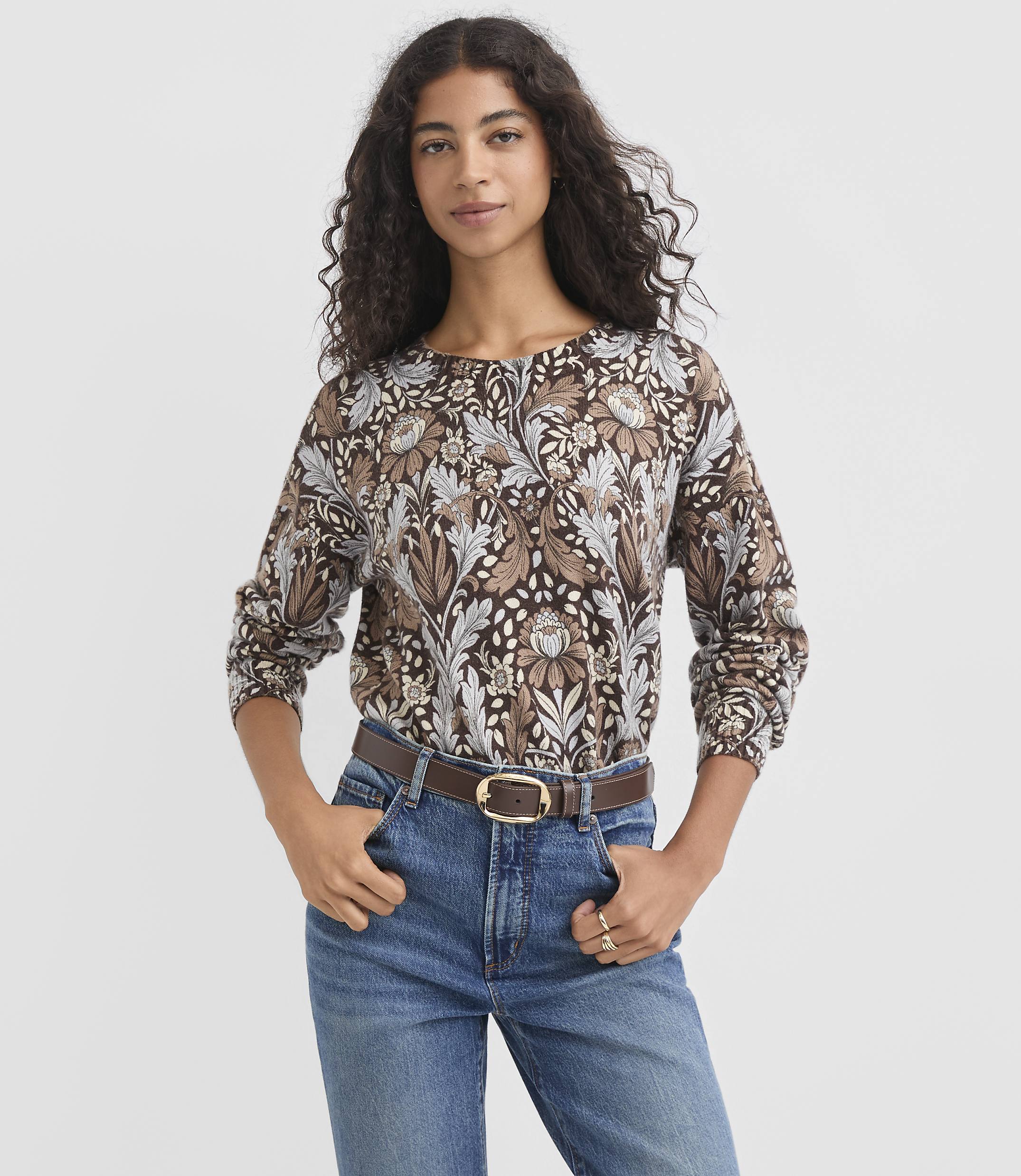 A woman in a patterned brown shirt and blue jeans stands against a plain gray background.