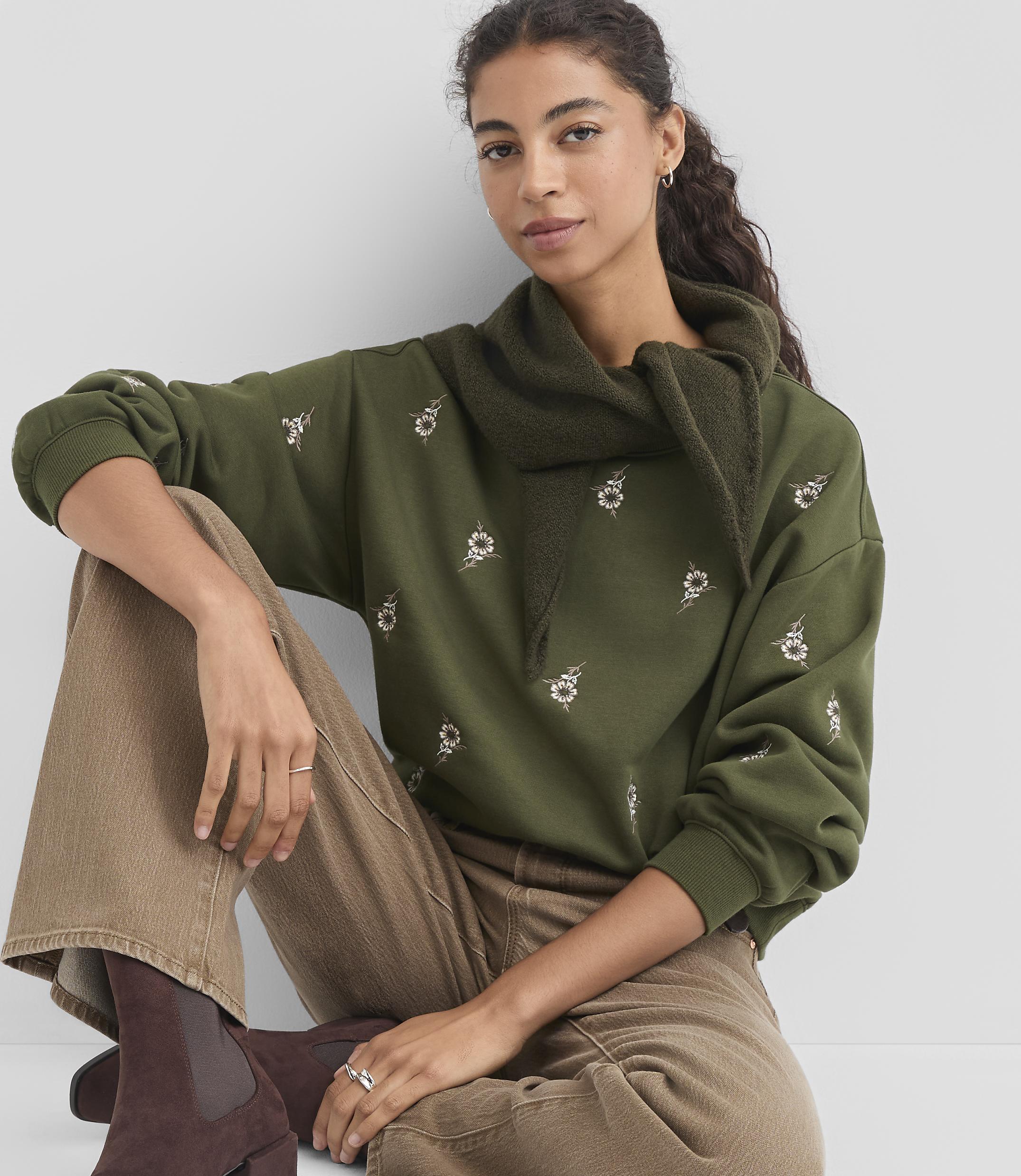A woman in a green embroidered sweatshirt and brown pants sits against a plain gray background.