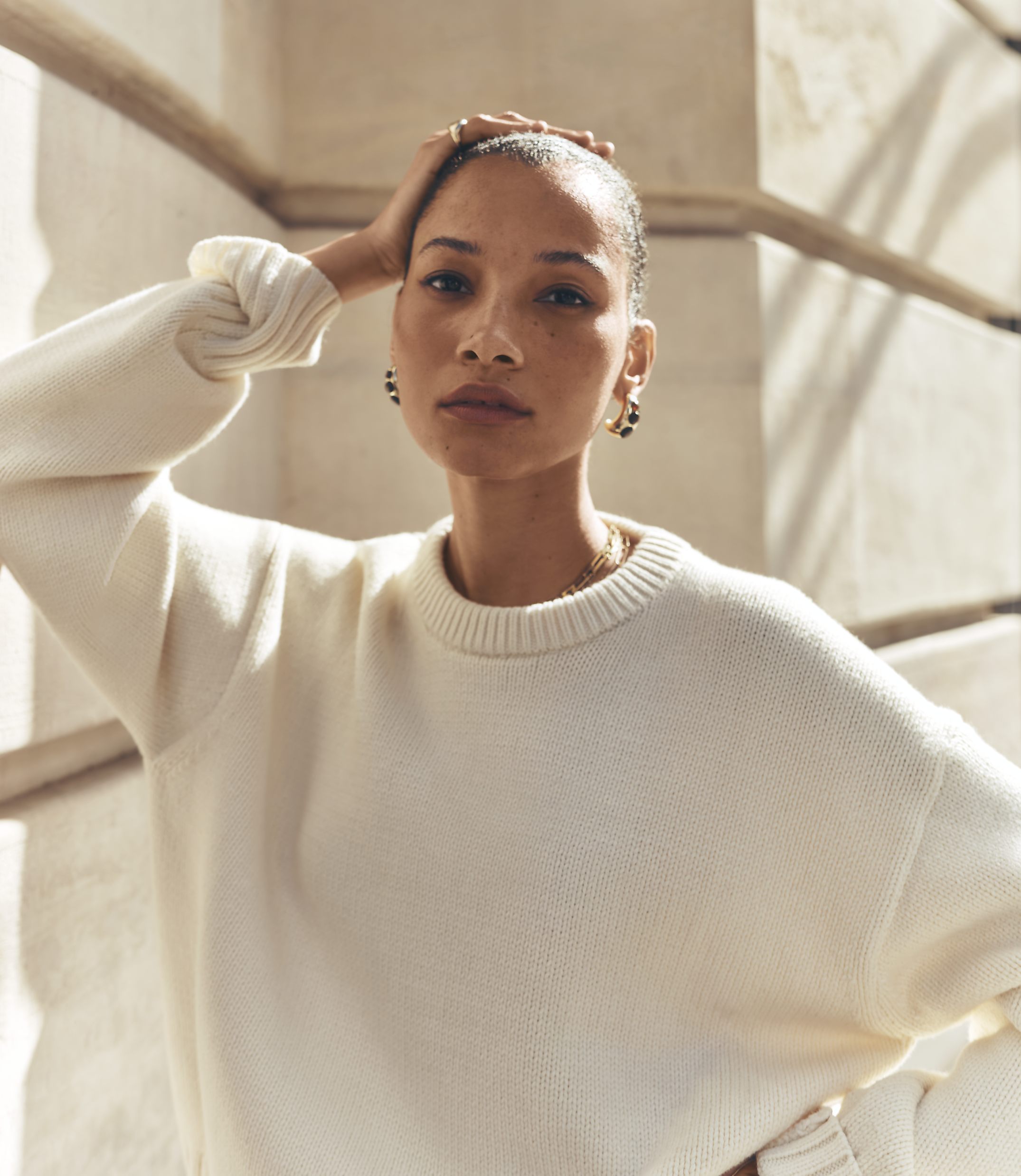 A woman in a cream sweater poses with her hand on her head against a blurred background of architectural details.
