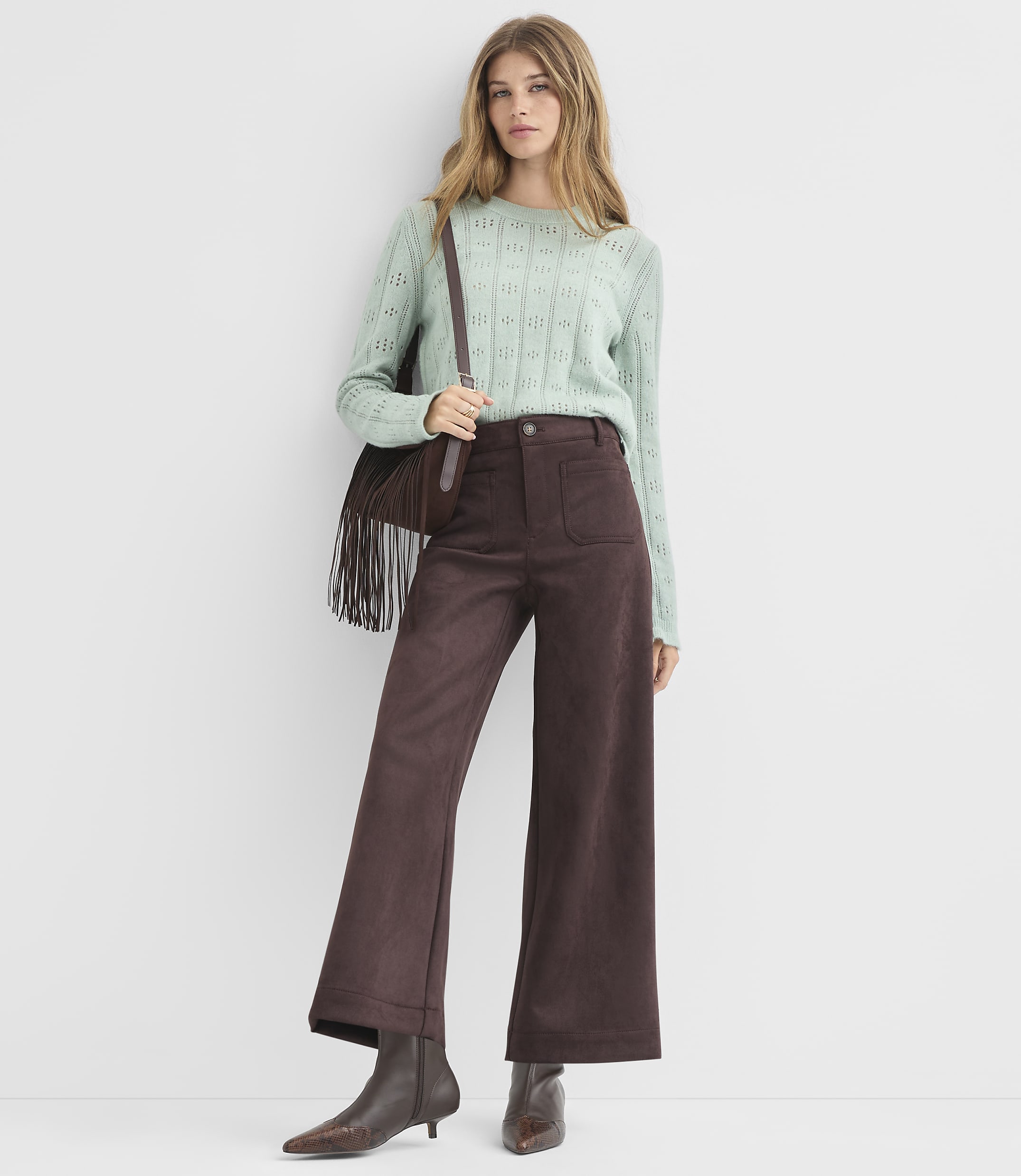 A woman in a light green sweater and brown wide-leg pants stands against a plain white background.