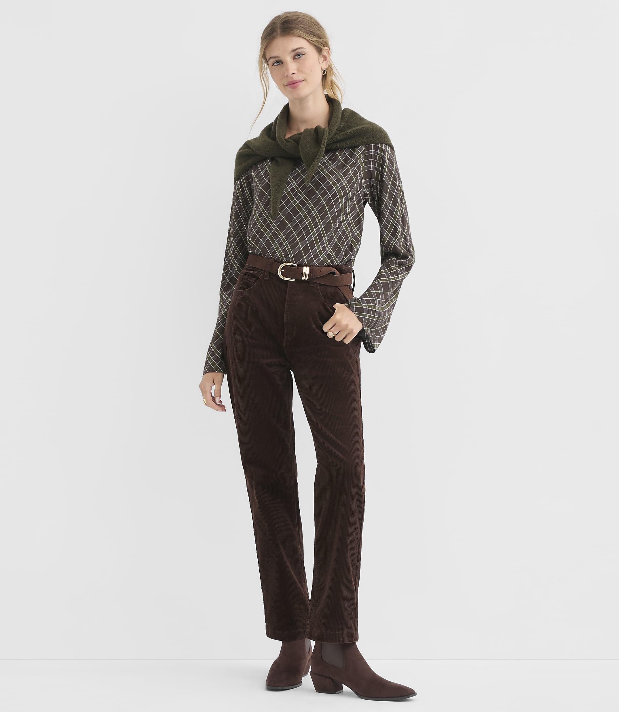 A woman in a brown plaid shirt, brown corduroy pants, and a green sweater draped over her shoulders stands against a plain white background.