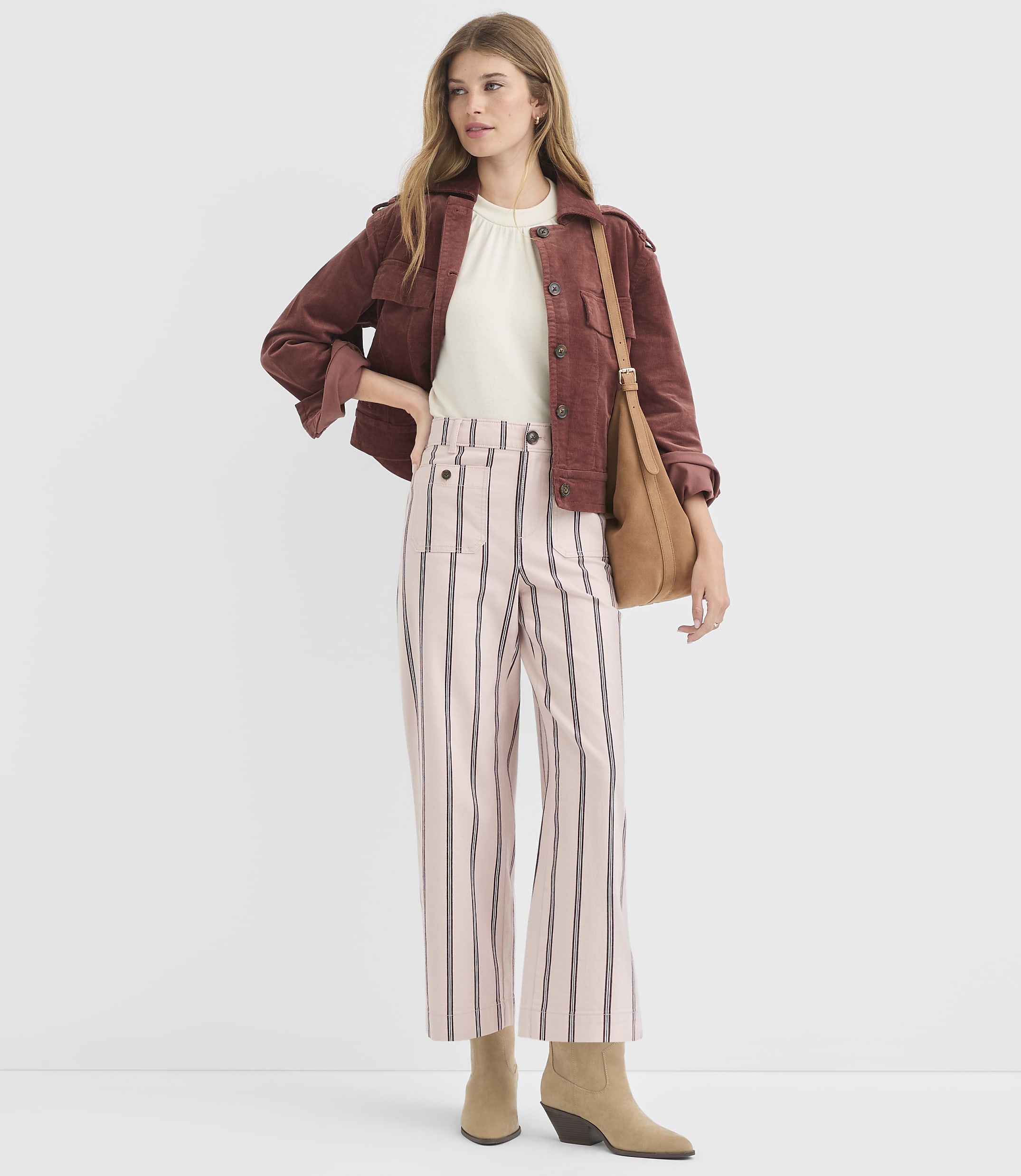 A woman in a maroon jacket and pink striped pants stands against a plain white background, holding a brown purse.
