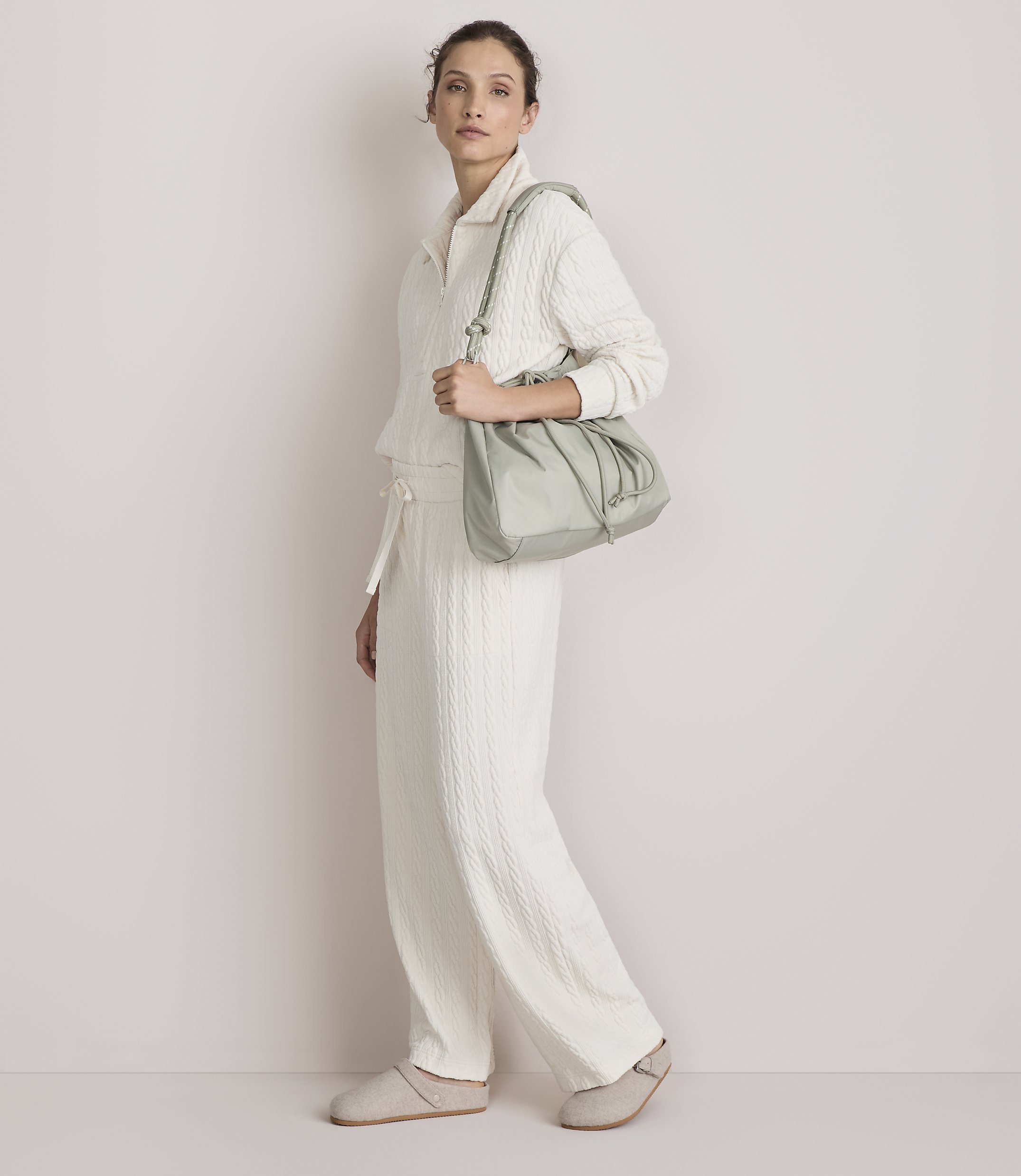 A woman in a cream-colored loungewear set and slippers walks with a light green drawstring bag against a plain, light-colored background.